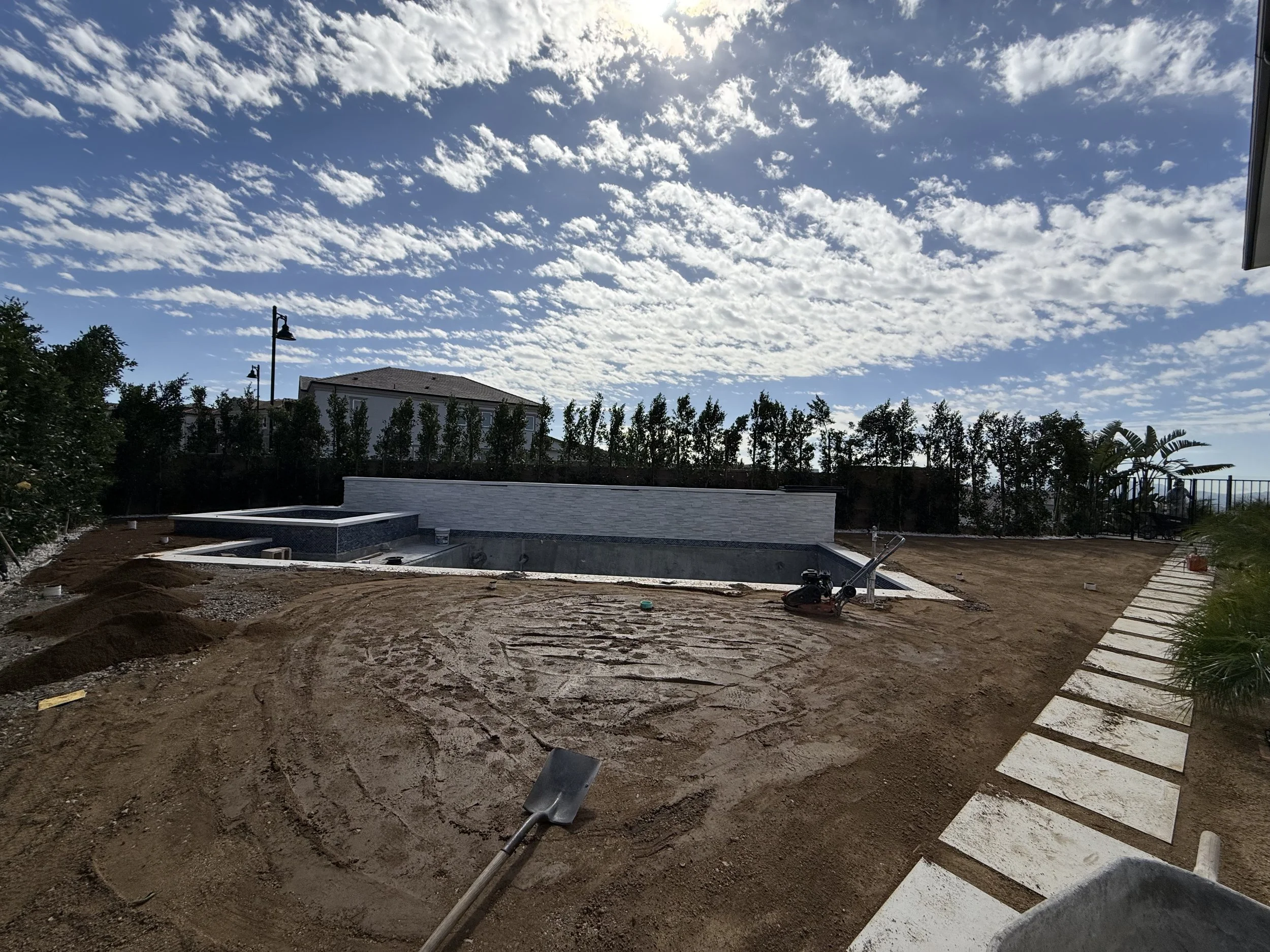 Unfinished backyard with a pool, surrounded by construction materials, dirt ground, and a paved walkway with white stones, with a house and trees in the background under a partly cloudy sky.