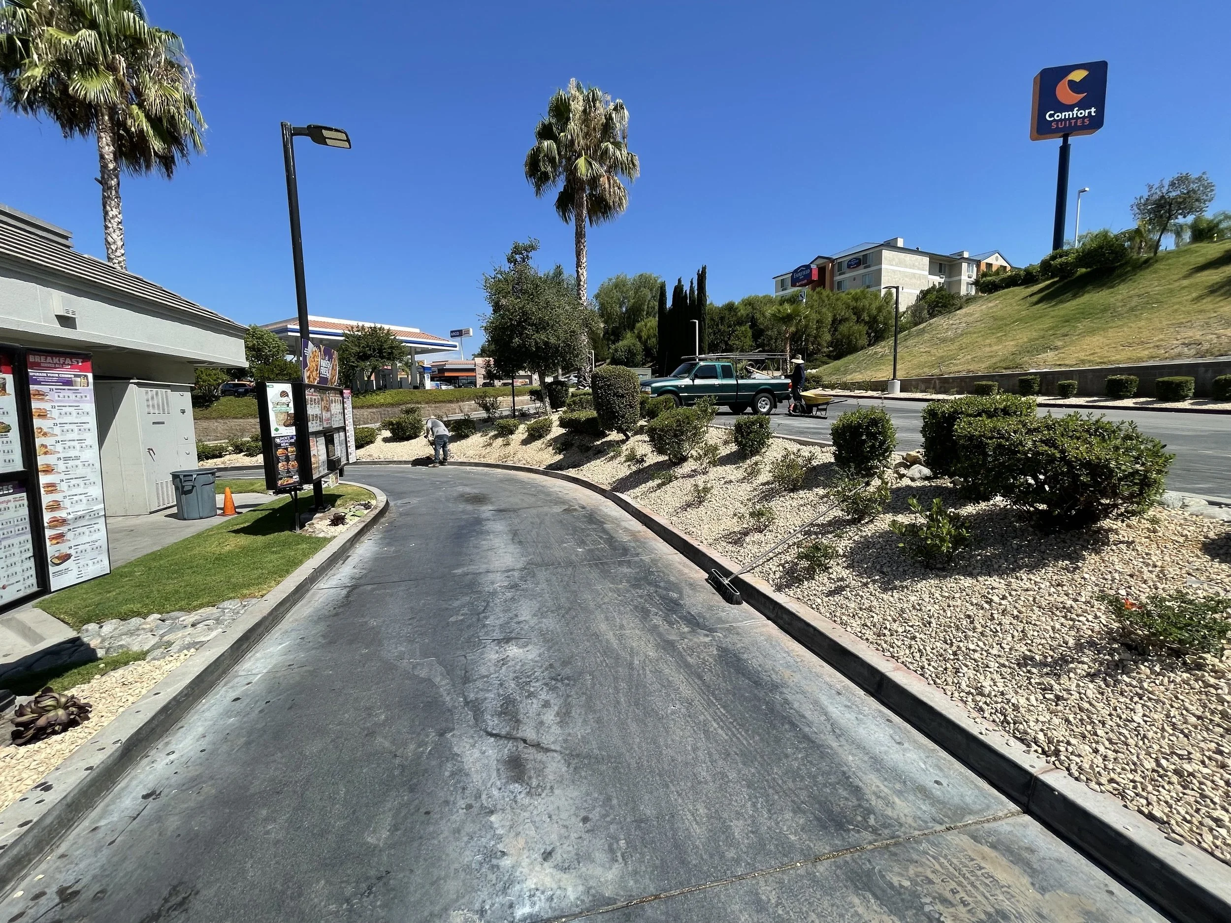 Fast food restaurant drive-thru with menu boards on the left, a green pickup truck parked in front, and a worker with a cart on the right. Tall palm trees and a Comfort Suites hotel building in the background under a clear blue sky.