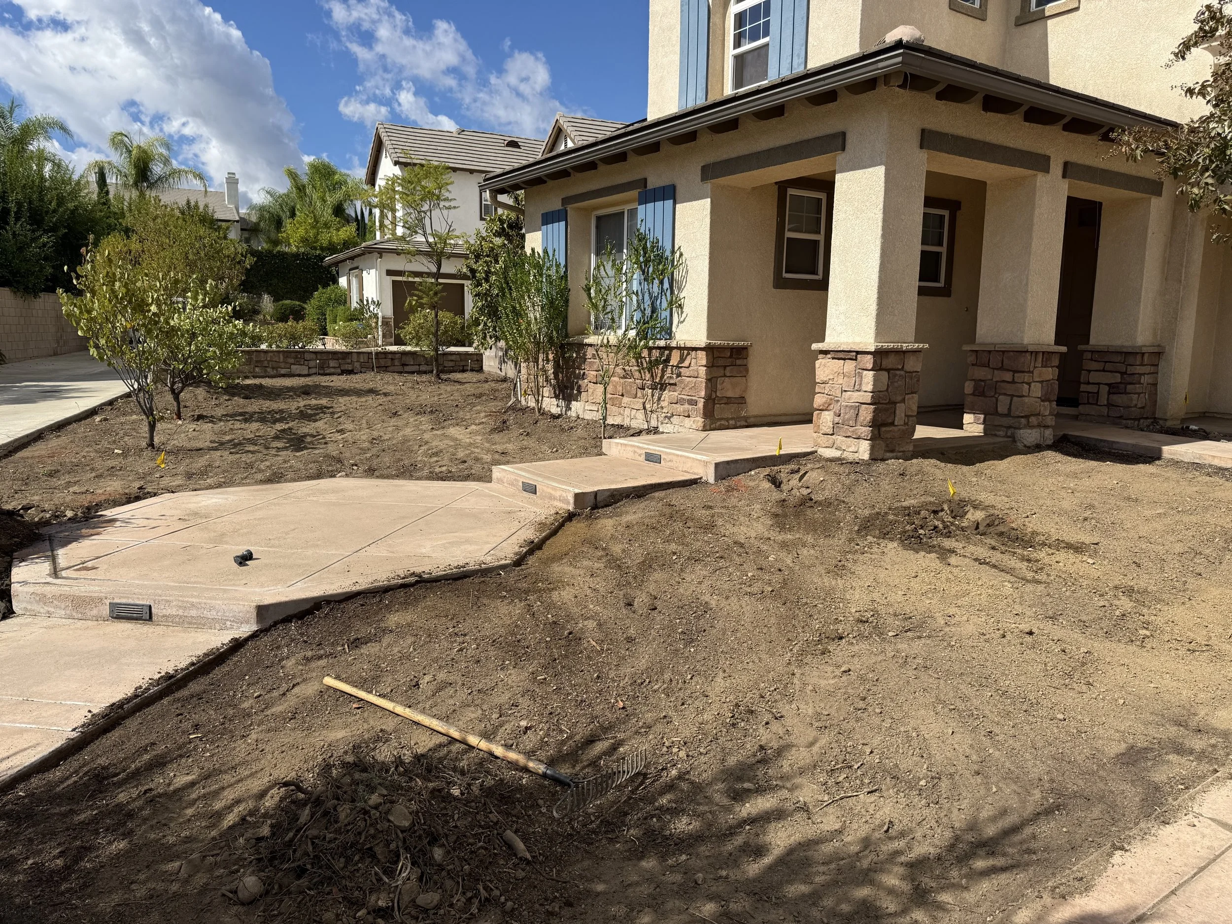 Front yard of a house under landscaping with new concrete sidewalk, small trees, and patch of dirt on a sunny day with blue sky and clouds.