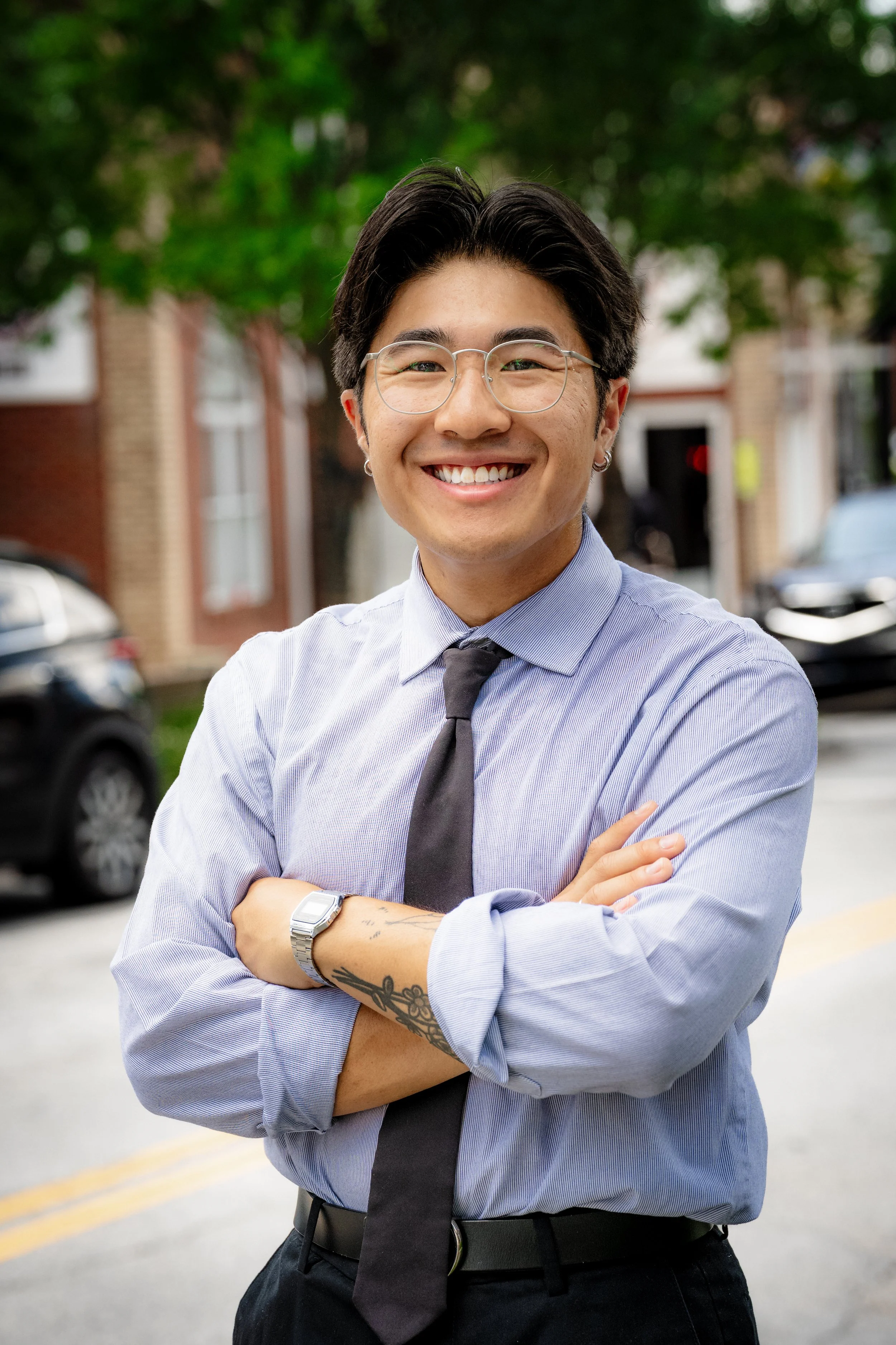 A young man standing outdoors smiling with arms crossed, wearing glasses, a light purple dress shirt, a black tie, and a silver watch, with a tree and parked cars in the background.