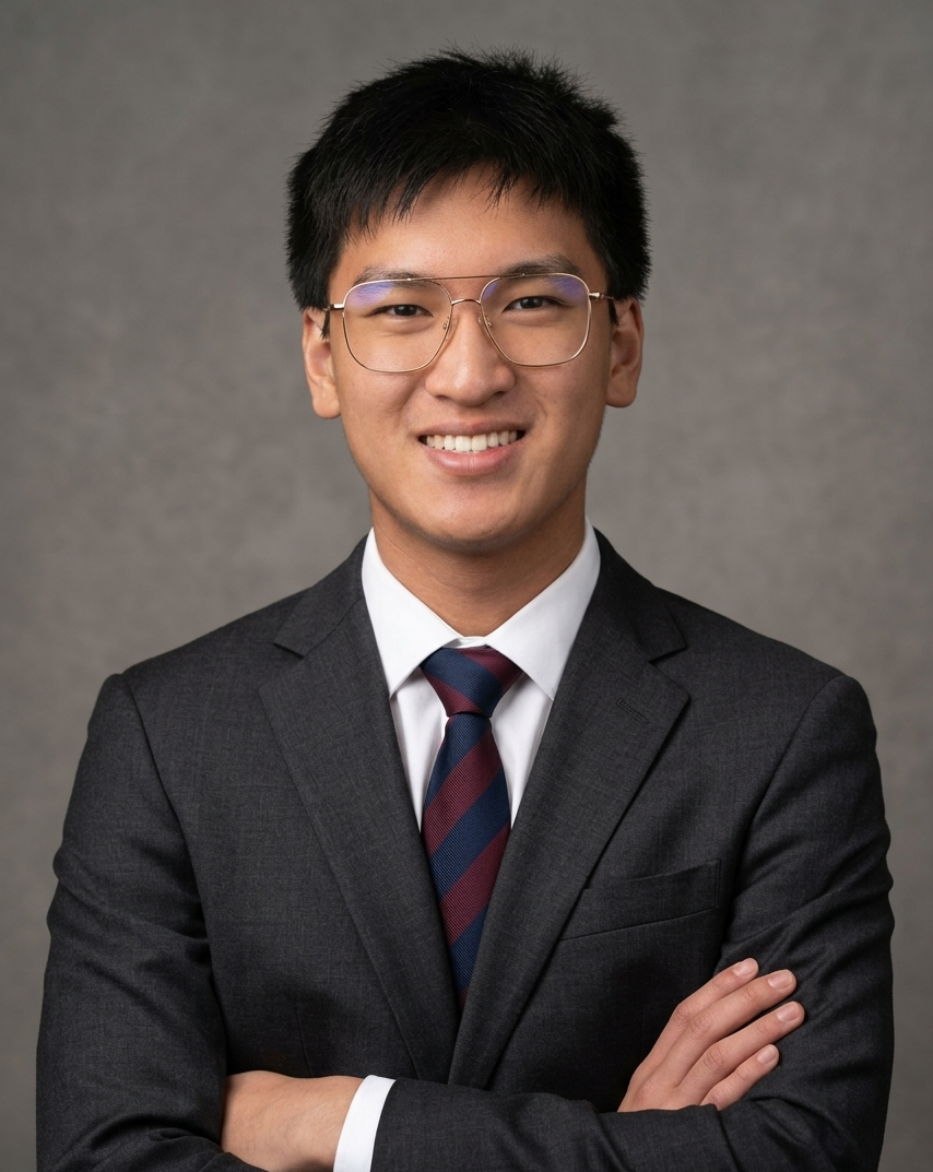 A young man wearing glasses, a dark gray suit, a white dress shirt, and a maroon and navy striped tie, standing with arms crossed and smiling in front of a gray background.