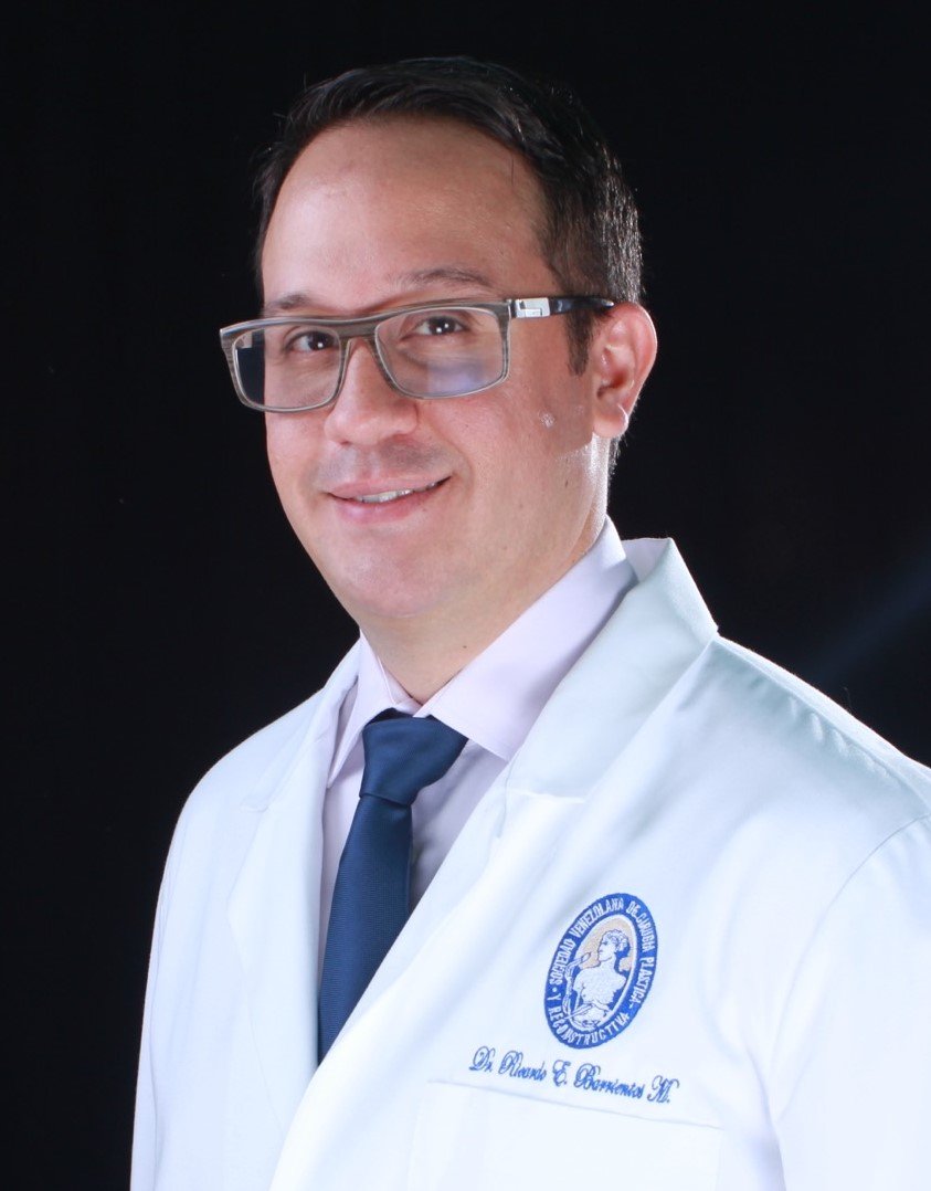 Professional portrait of a male doctor wearing glasses, a white lab coat with an embroidered logo, a shirt, and a tie, against a black background.