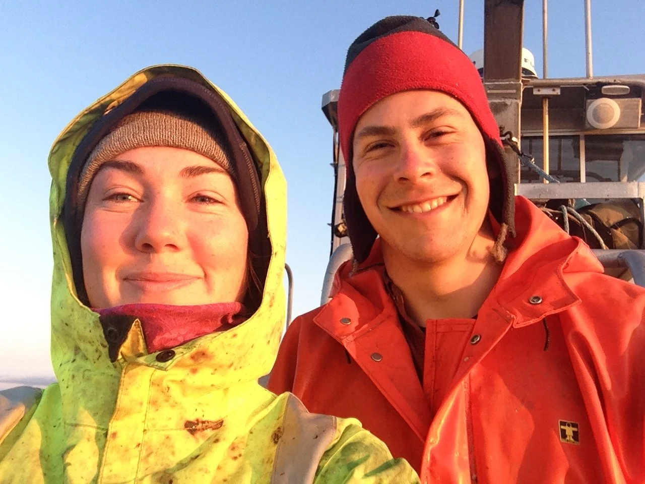Mariah and Jake, wearing their hard weather gear on their boat, taking a selfie outside with a clear sky in the background.