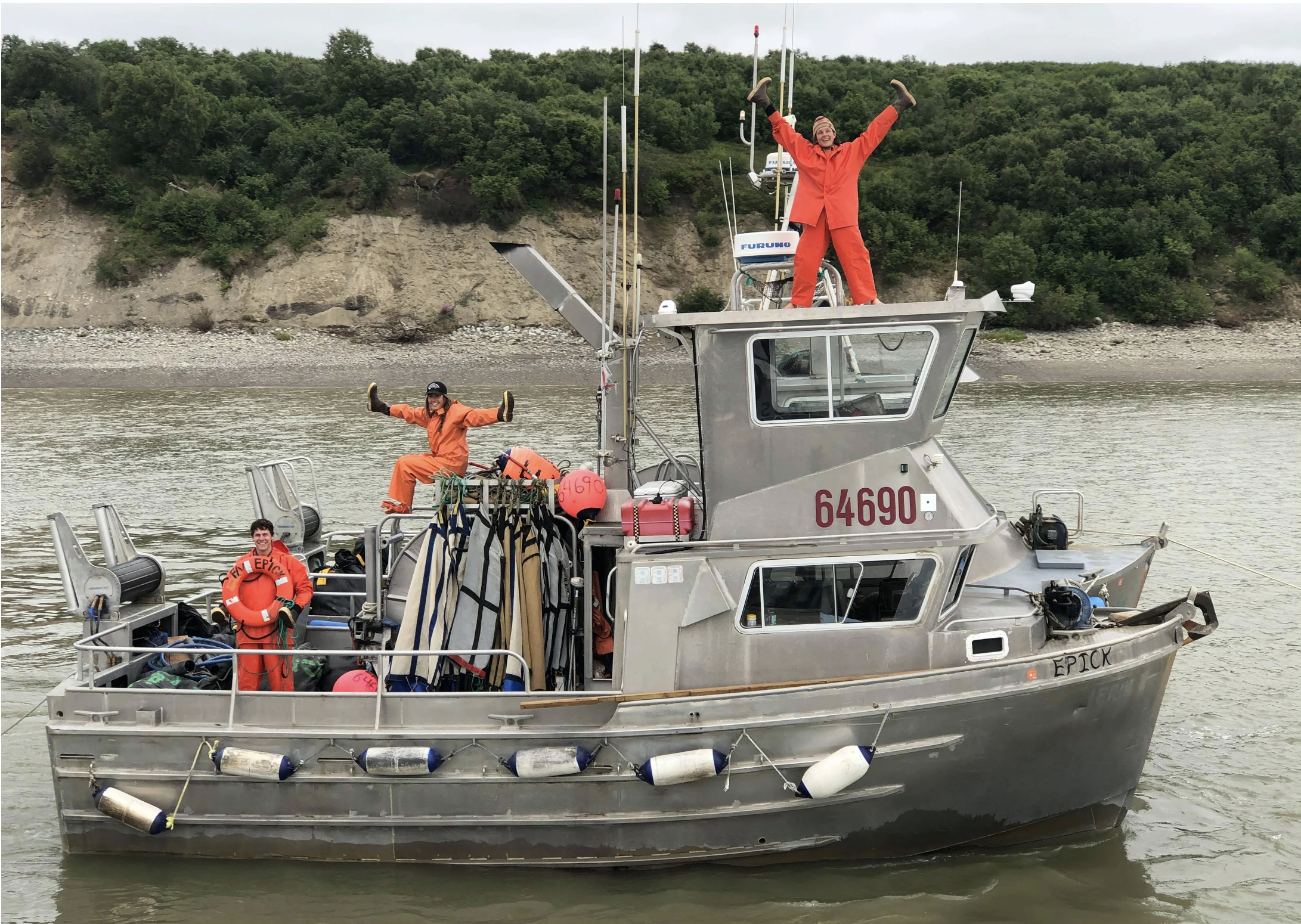 People on a small gray boat wearing orange life jackets and orange waterproof gear, smiling and waving, with a scenic river and green hillside in the background.