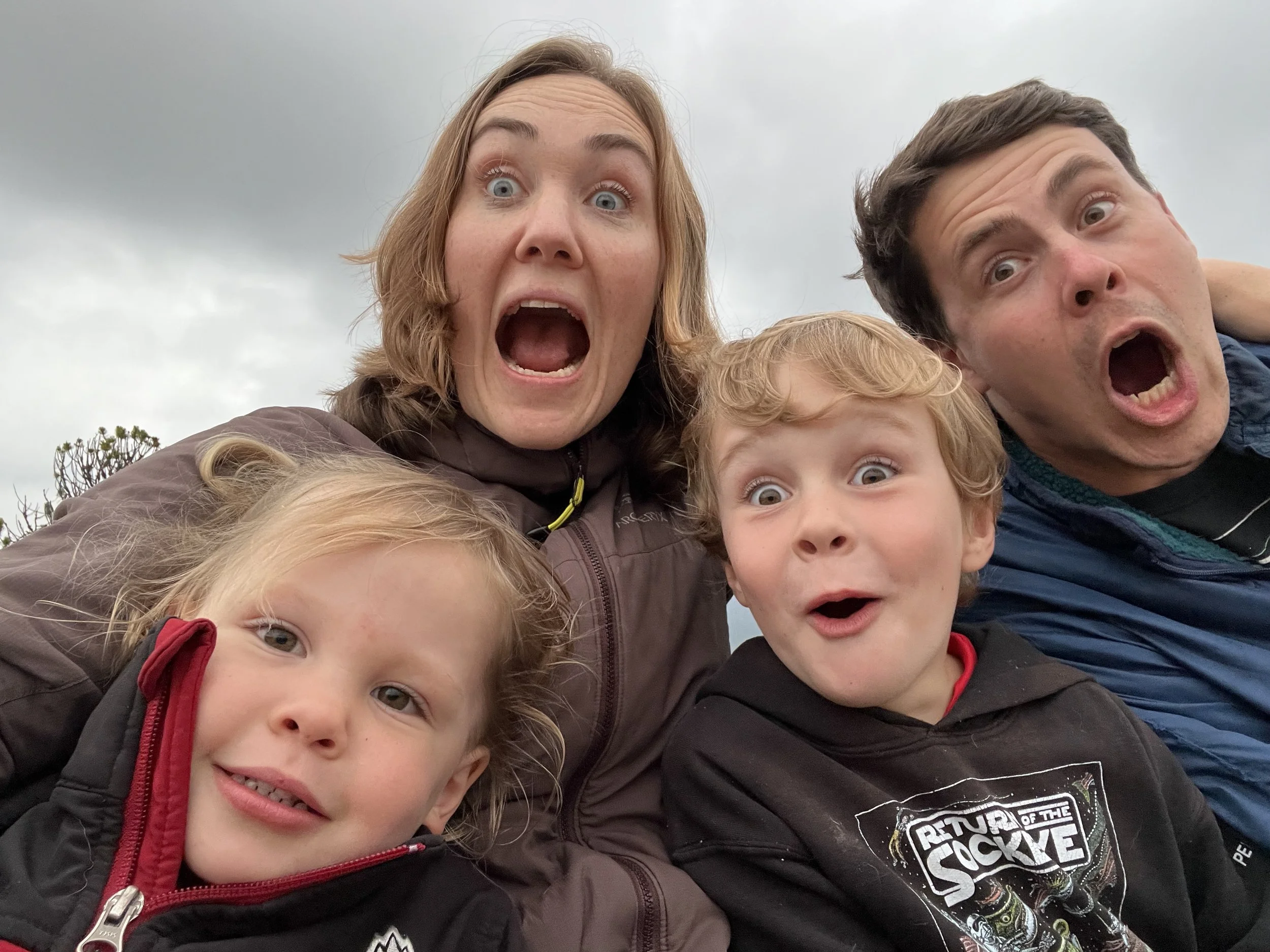 A group of four people, including two children and two adults, looking into the camera with surprised or excited expressions, outdoors on a cloudy day.