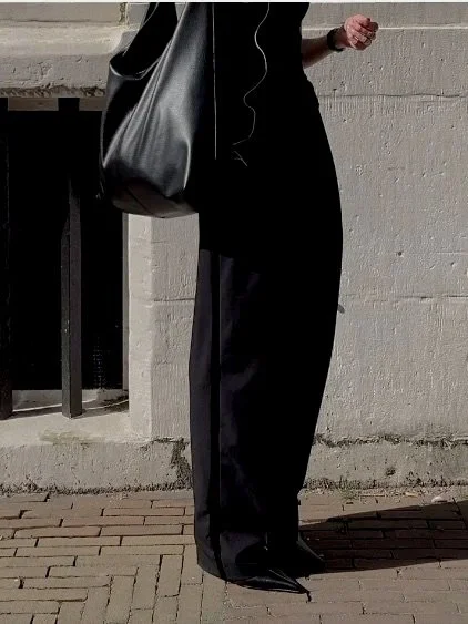 Person standing outdoors, wearing black pants and a long-sleeved top, holding a black tote bag and listening to headphones.