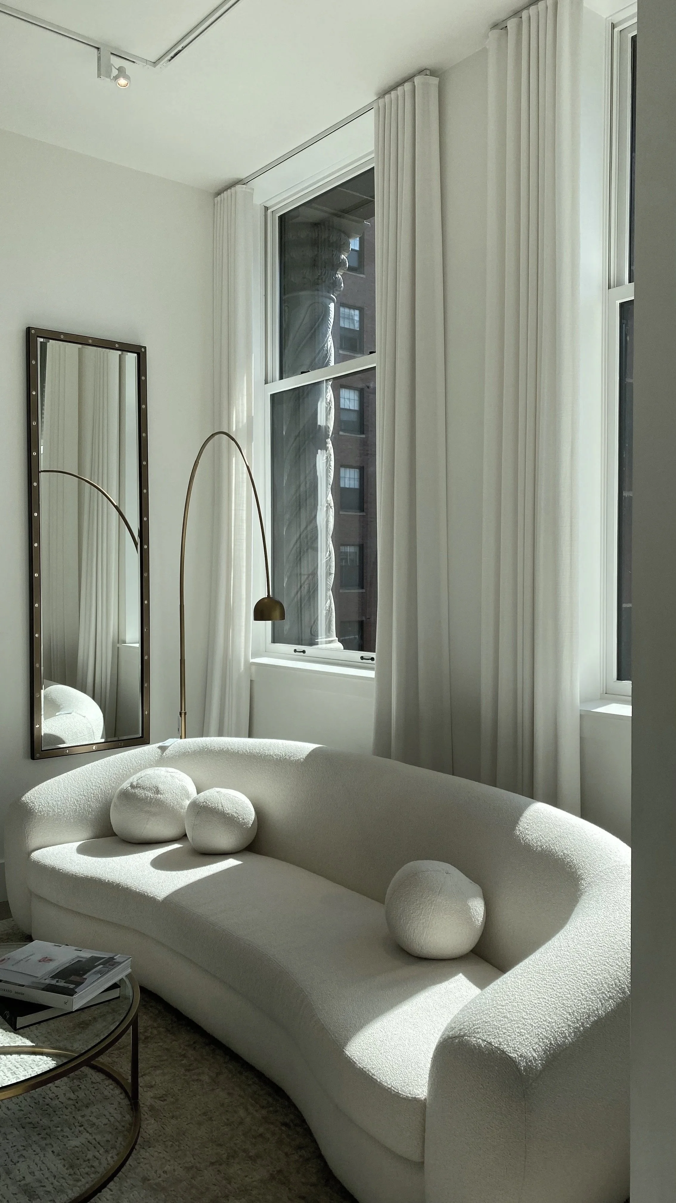 A modern, curved white sofa with spherical cushions in a bright, minimalist living room. There is a large window with white curtains, a mirror with a metallic frame, and a gold floor lamp. A book and a small round table are visible in the foreground.