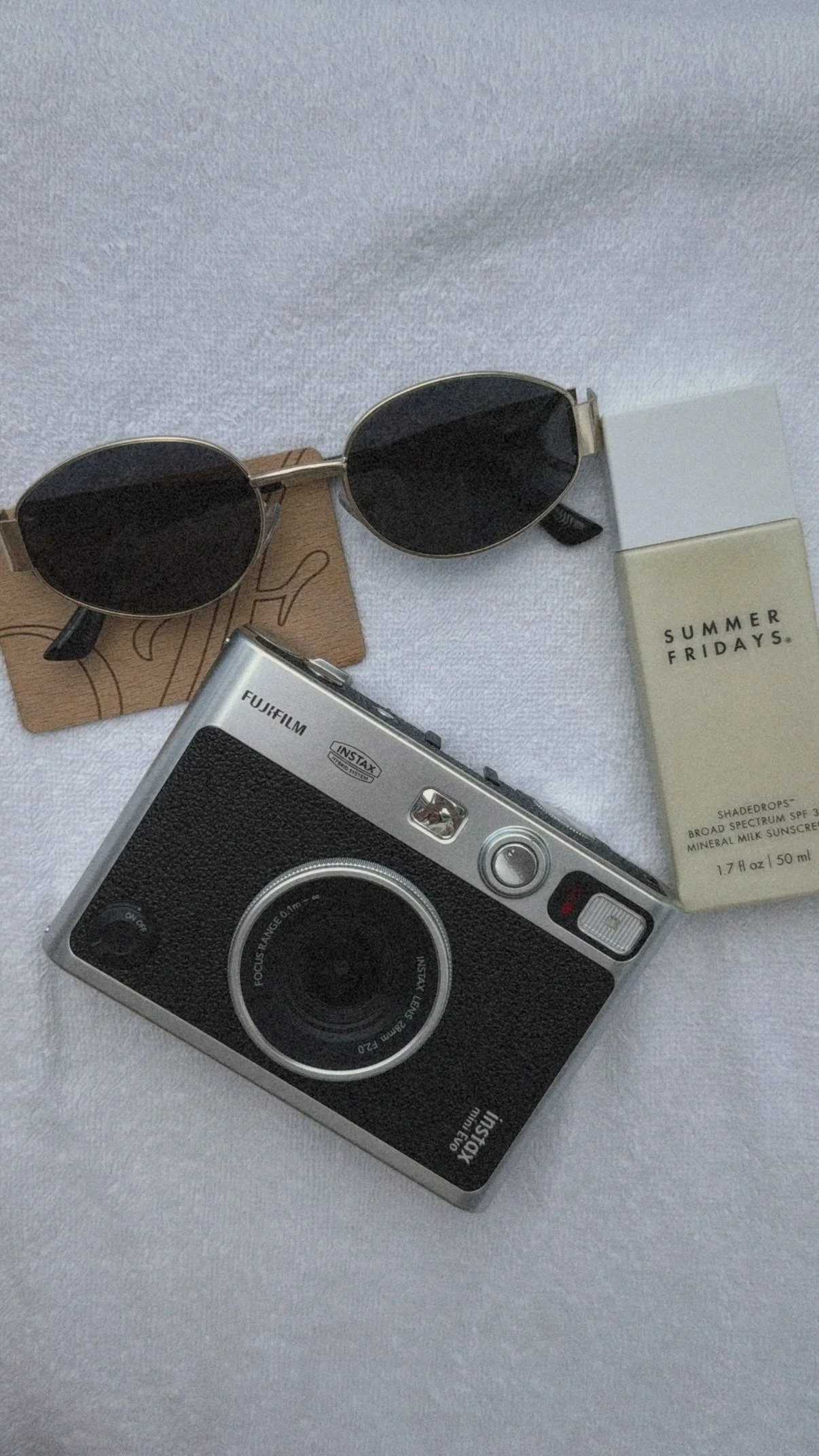 A pair of black sunglasses with gold frames, a vintage Fujifilm Instax instant camera, a small white box labeled "Summer Fridays" broad spectrum SPF 30 sunscreen, and a brown paper card with a line drawing of a face, all placed on a white textured surface.