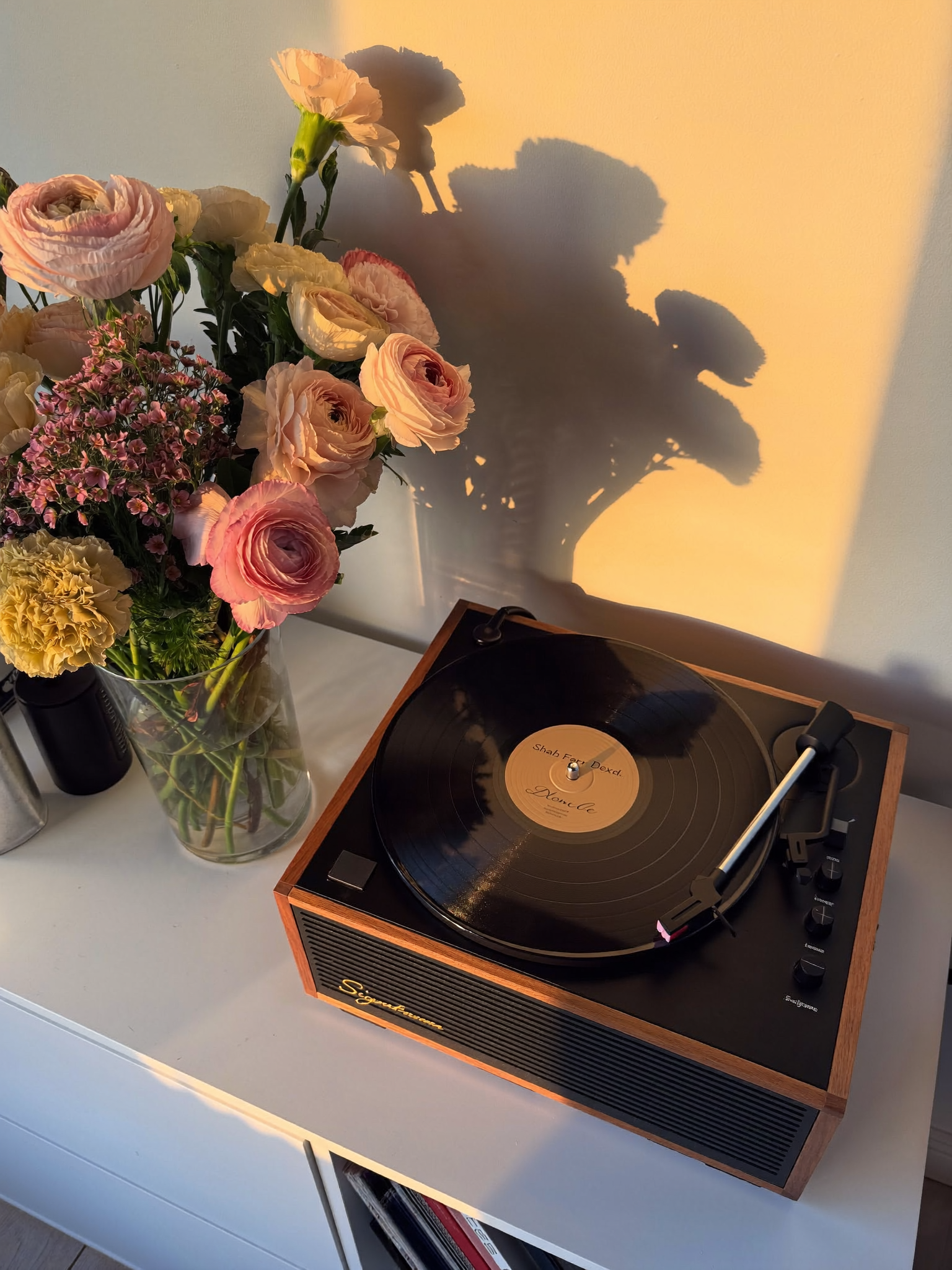 A vase of pink and cream flowers sitting on a white table next to a vintage record player, with sunlight casting shadows of the flowers on the wall.