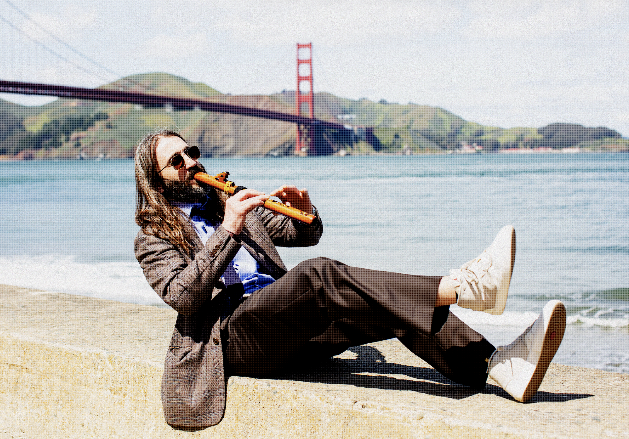 A man with long hair and a beard, wearing sunglasses, a brown checked blazer, and light-colored shoes, is sitting on a lounge chair at the beach, playing a yellow flute. In the background, the Golden Gate Bridge is visible over a body of water with rolling green hills.