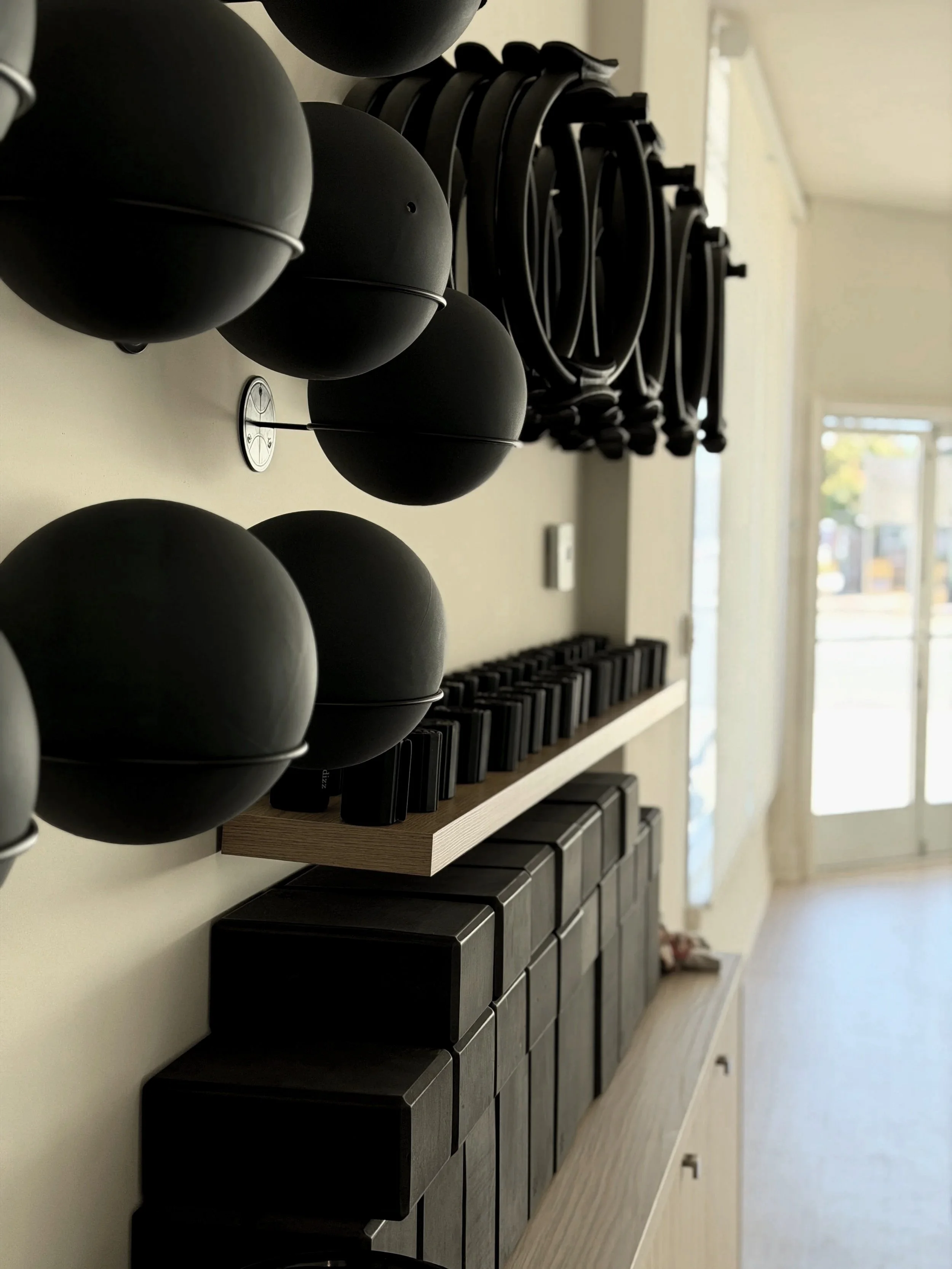Pilates & Yoga balls, rings, and blocks are mounted on a wall above on a wooden shelf in a well-lit room.
