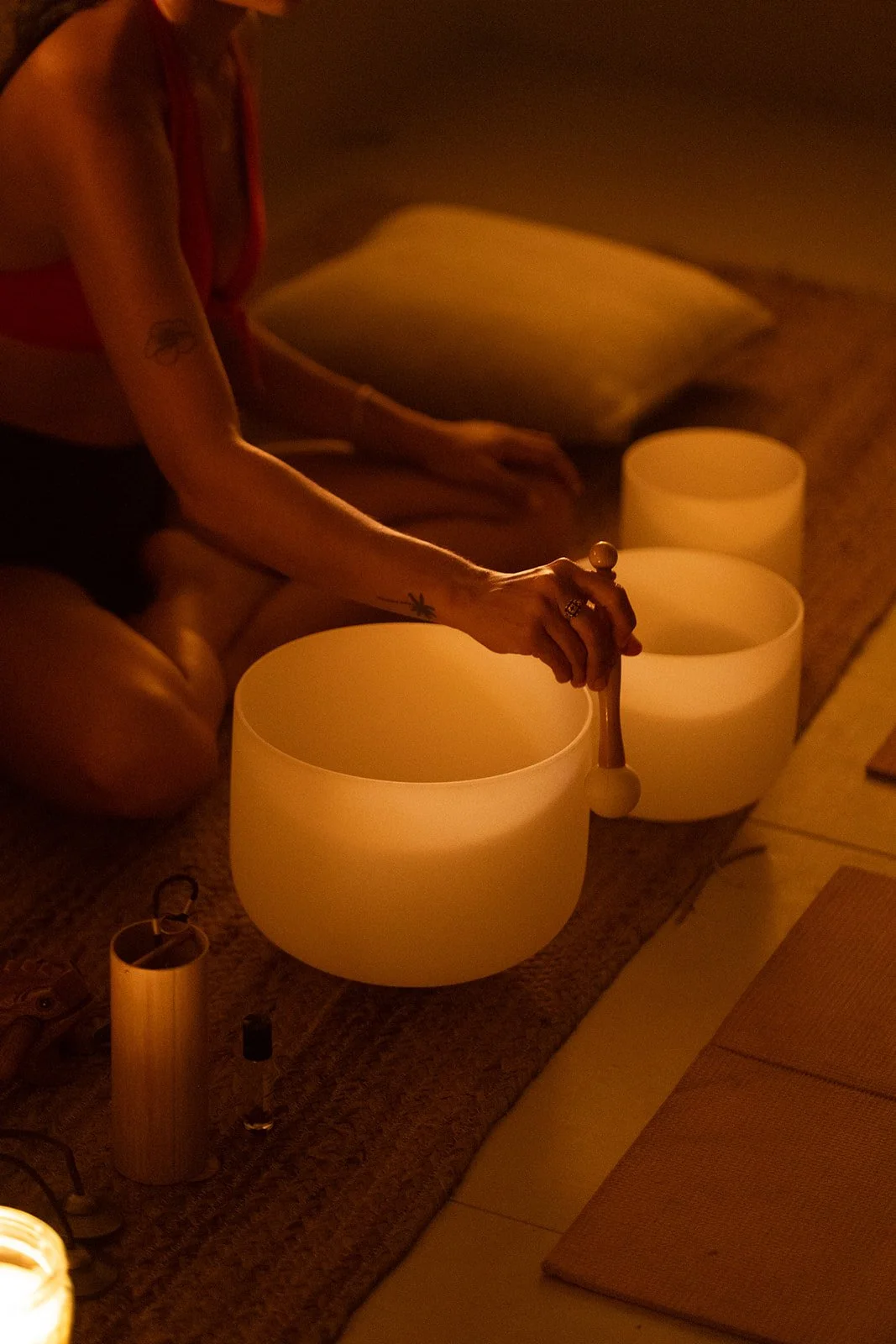 Person playing crystal singing bowls in a softly lit room with warm lighting, sitting on a floor with cushions.
