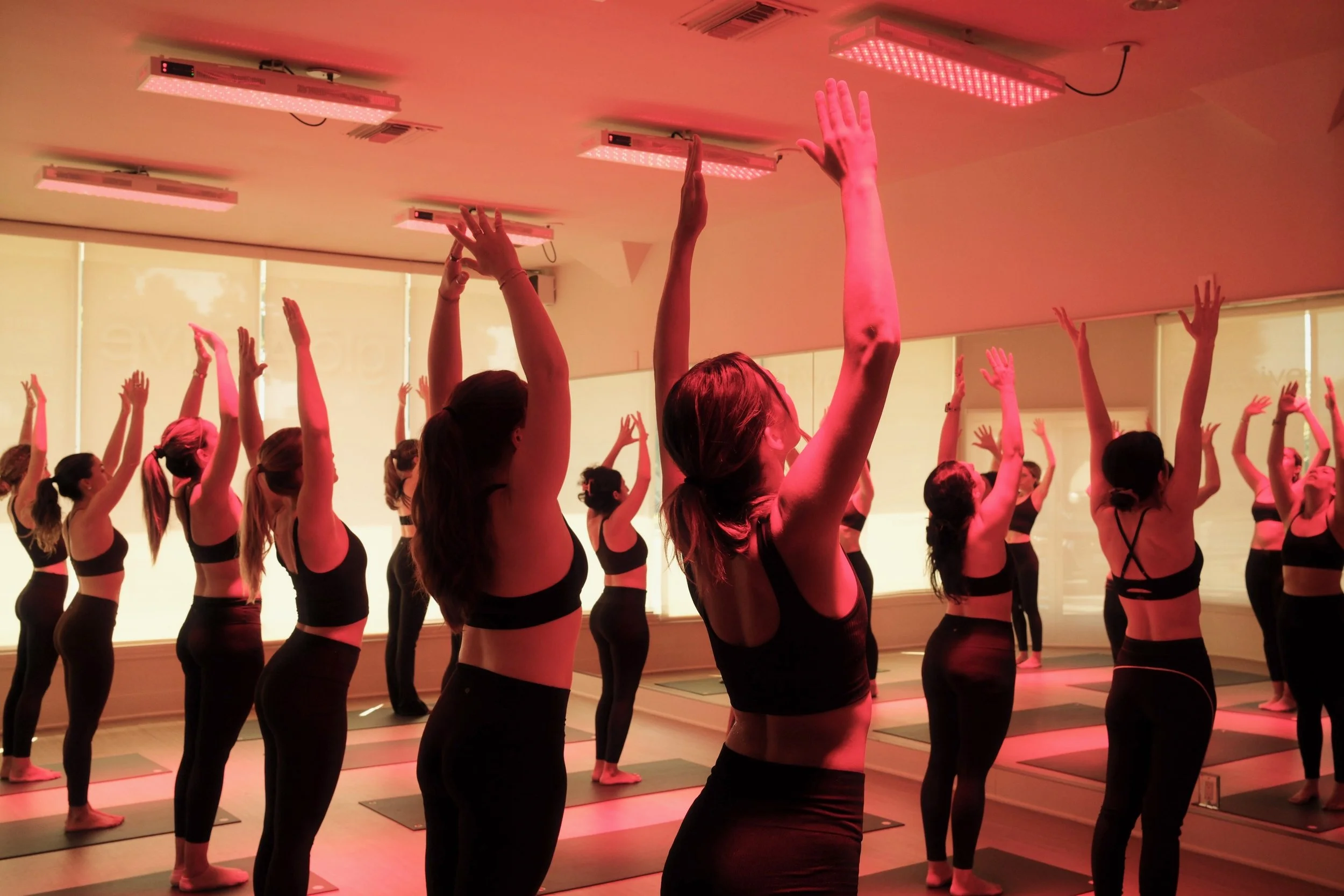 A group of women participating in a yoga or fitness class in a studio, standing on yoga mats with their arms raised, illuminated by red light therapy from ceiling-mounted LED lights.