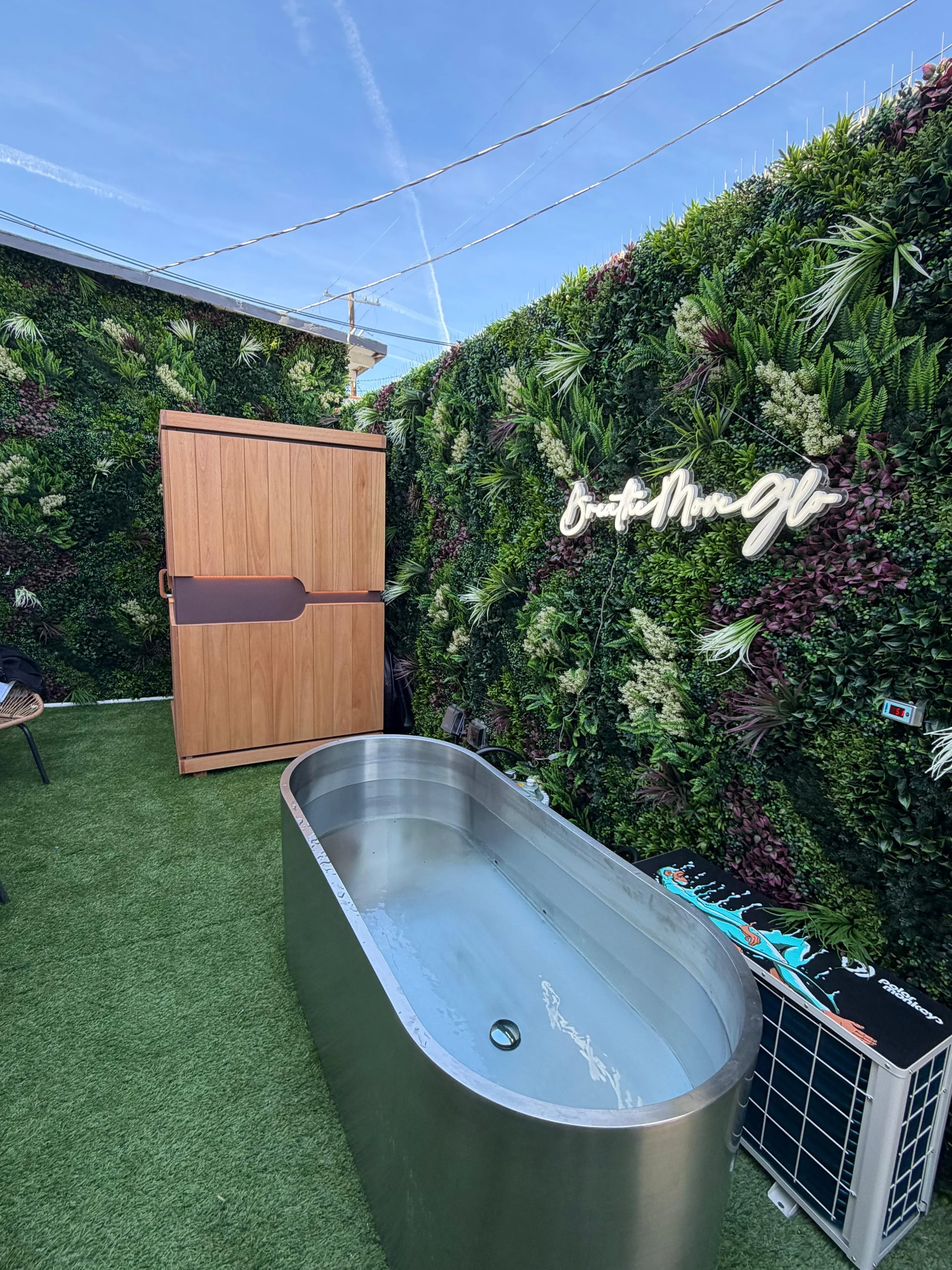 Cold Plunge Tub on green grass next to a wall covered with green plants and a neon sign that says 'Breathe. Move, glō' under a clear blue sky. A wooden infrared sauna is seen to the left of the cold plunge tub.