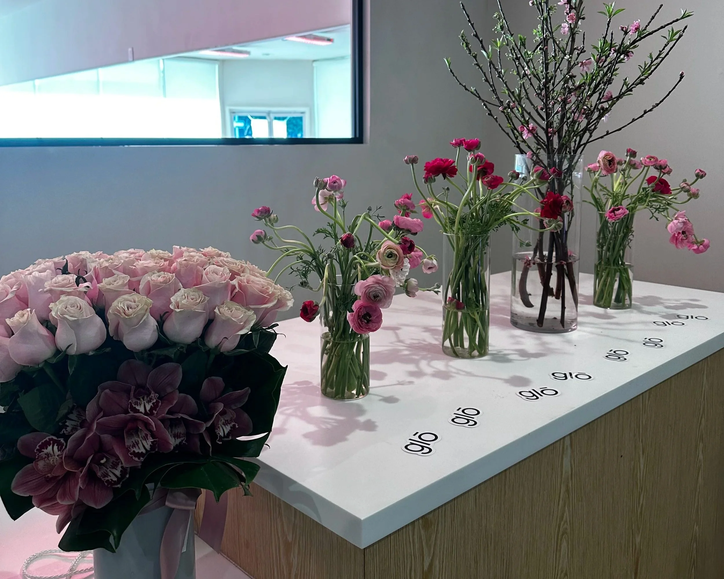 Several vases filled with pink and purple flowers displayed on a white table, with a window in the background showing a studio setting.