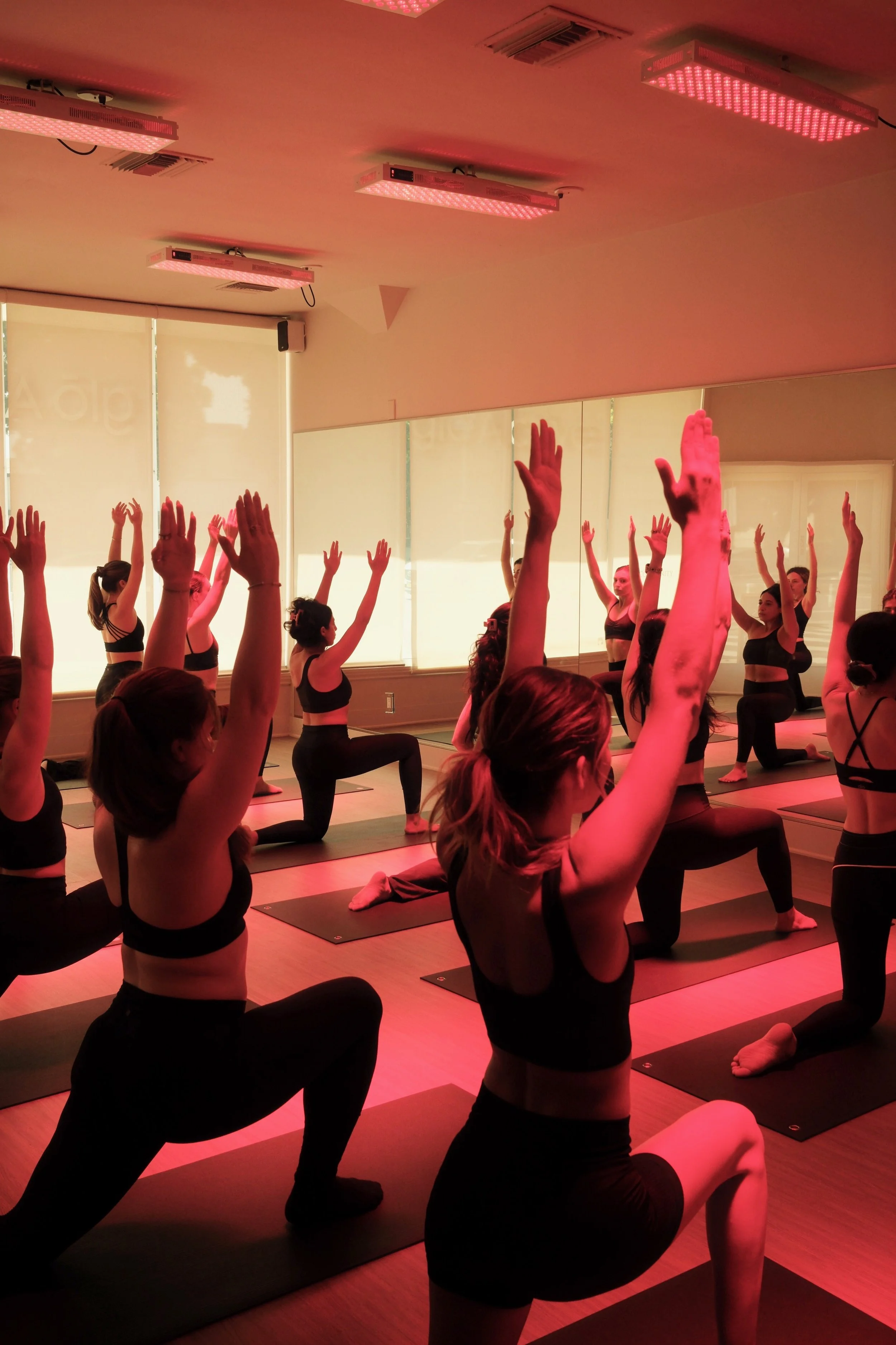 A group of women practicing yoga in a dimly lit studio with red light therapy. They are performing a kneeling pose with one leg extended back and both hands raised overhead. The studio has large windows with blinds and a mirror along the wall.