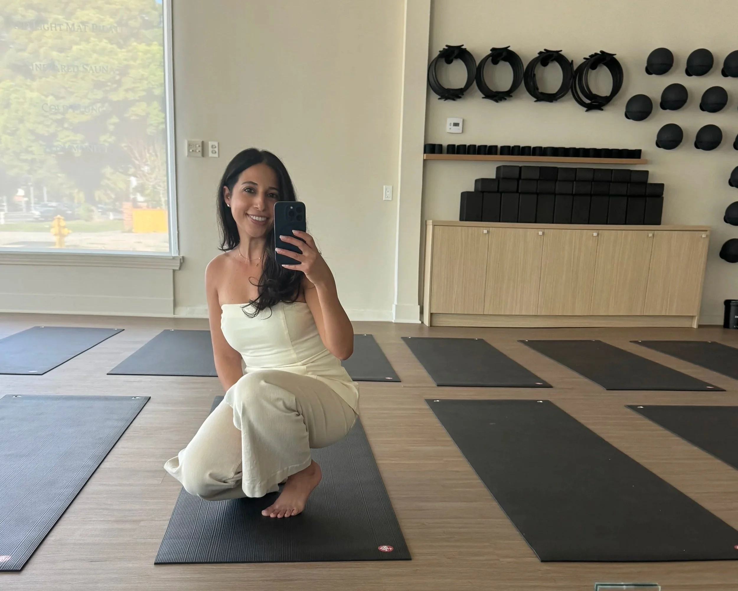 The female owner of glō active in a cream-colored outfit taking a mirror selfie in a yoga studio with yoga mats and equipment on the wall.