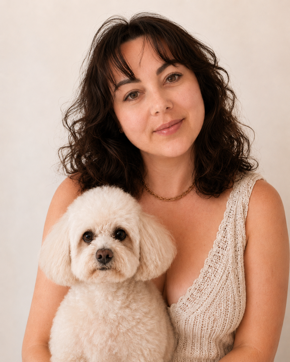 Eine Frau mit braunen, lockigen Haaren und ein cremefarbenes Top hält einen weißen Hund vor neutralem Hintergrund.