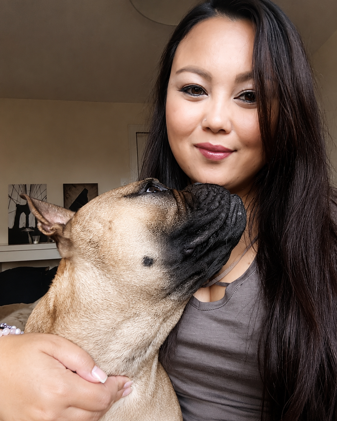 Eine Frau mit langen dunklen Haaren hält einen French Bulldog, der den Kopf an ihr Gesicht reibt, in einem Wohnzimmer.
