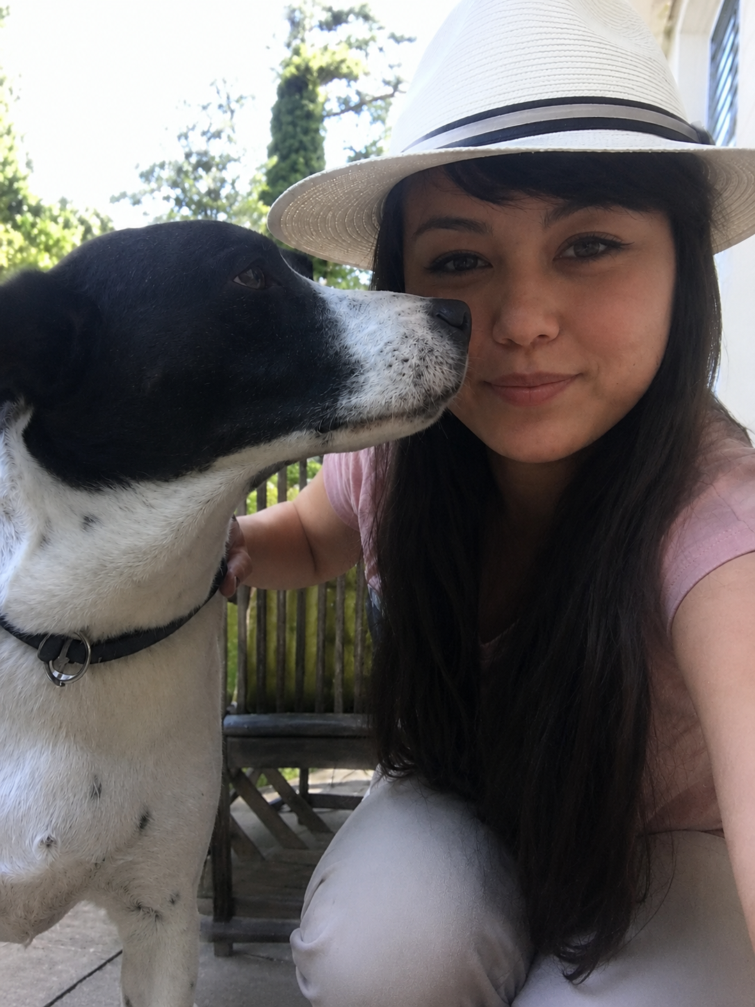 Eine junge Frau mit braunen Haaren, weißer Hut und rosa Shirt macht ein Selfie mit einem schwarzen und weißen Hund im Freien, im Hintergrund Bäume.