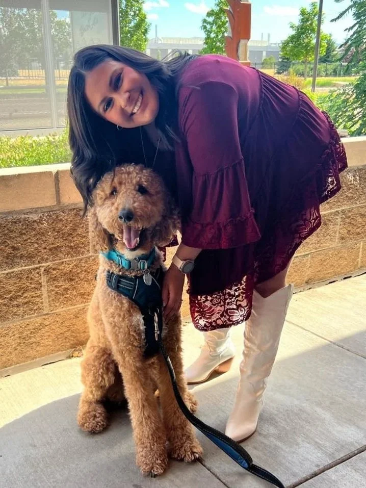 A woman in a burgundy dress and white boots is smiling and leaning towards a brown poodle wearing a harness, outside on a sidewalk near a brick wall and a window, with trees and a building in the background.