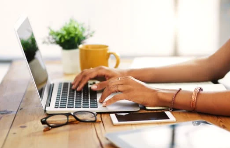 Person typing on a laptop with a coffee mug, glasses, tablet, and phone on a wooden desk.