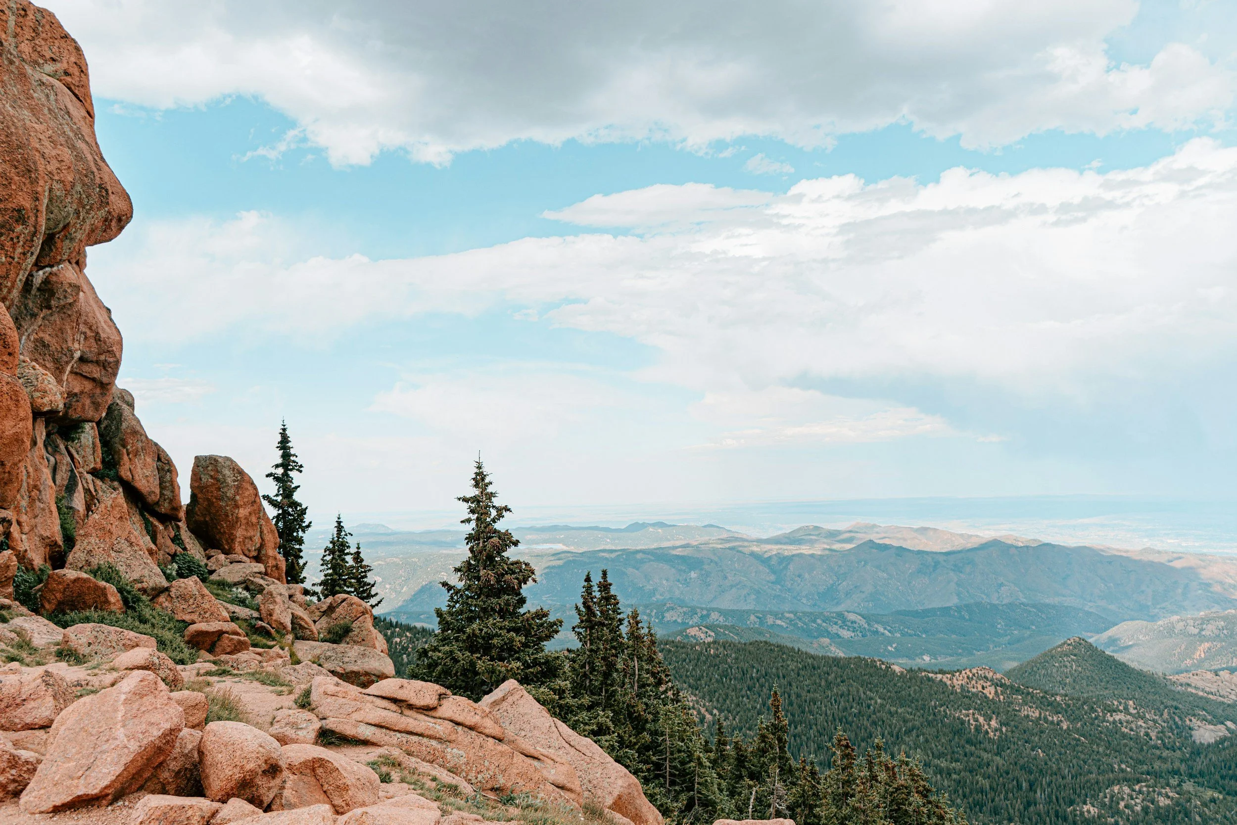 Rocky mountain landscape with large reddish-brown rocks in the foreground, pine trees scattered across the middle ground, and layered mountain ranges extending into the distance under a partly cloudy sky.