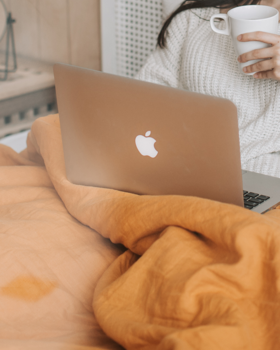 A person wearing a white knitted sweater holding a white mug, with a silver Apple laptop resting on an orange blanket.