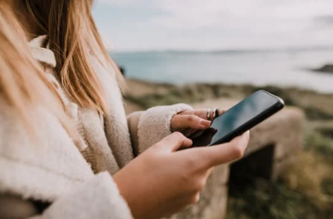 Person holding smartphone outdoors near a coastal area with rocks and water in the background.