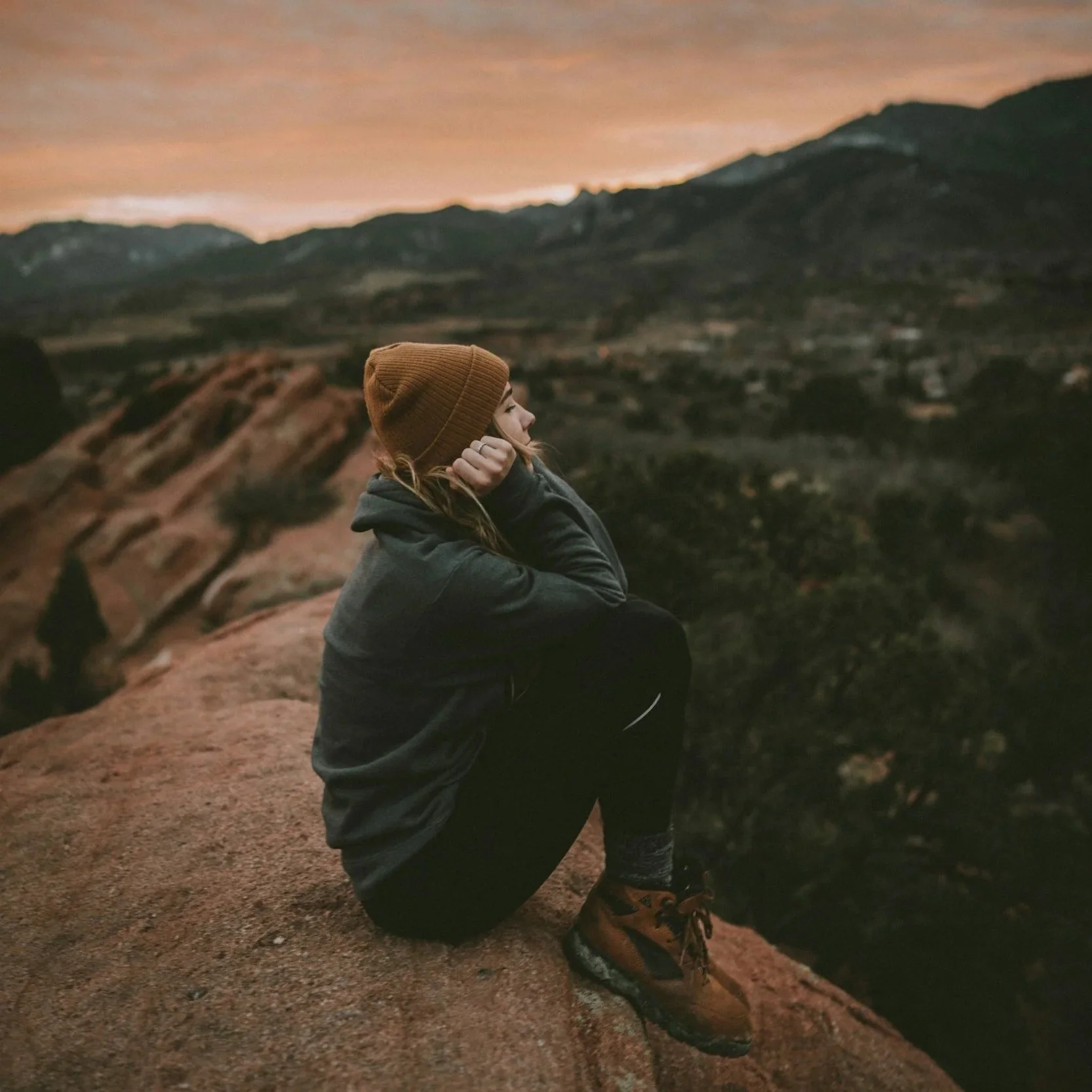 A person sitting on a rocky terrain at sunset, wearing a brown beanie, gray hoodie, and orange hiking boots, with mountains and an orange sky in the background.