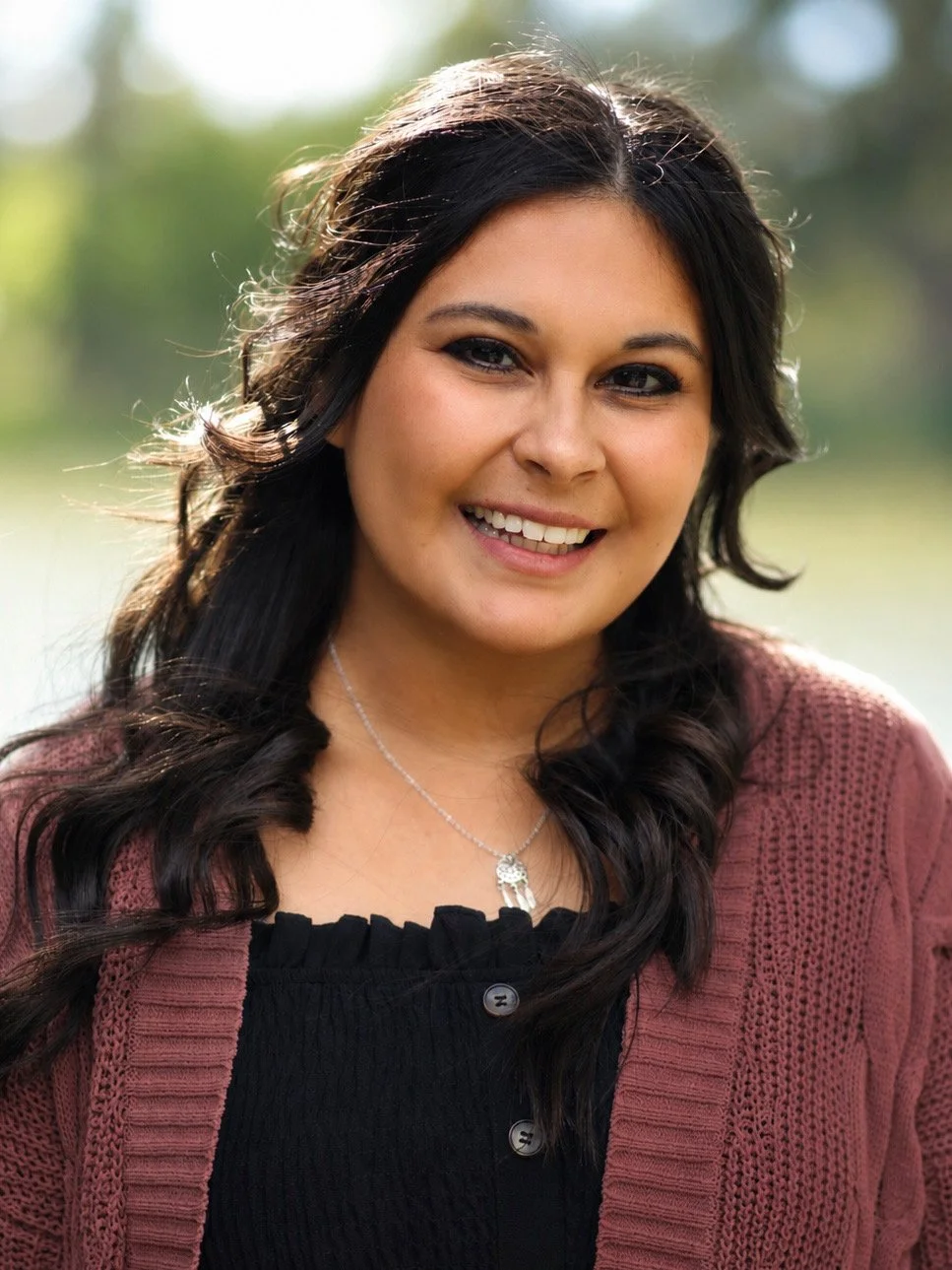 A woman with long dark hair smiling outdoors with a blurred green background, wearing a pink cardigan over a black top and a silver necklace.