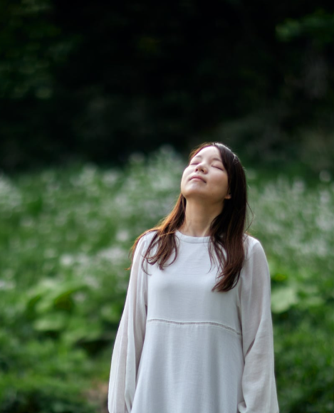 Woman with brown hair wearing a white long-sleeve shirt, standing outdoors with eyes closed, in a lush green environment.