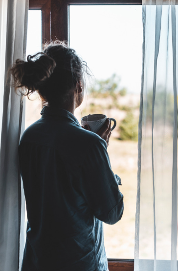 A woman standing by a window, holding a mug and looking outside