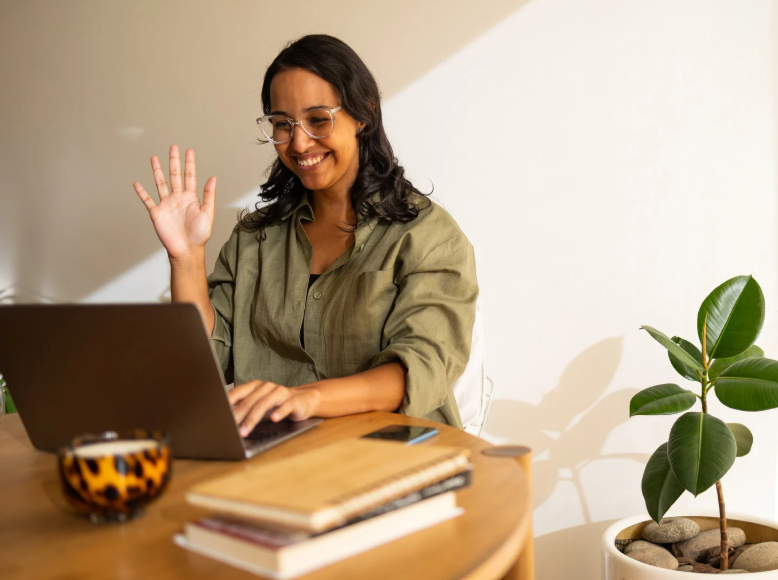 Woman with glasses, smiling and waving while working on a laptop at a wooden desk with books, a smartphone, and a mug, in front of a white wall with a potted plant.
