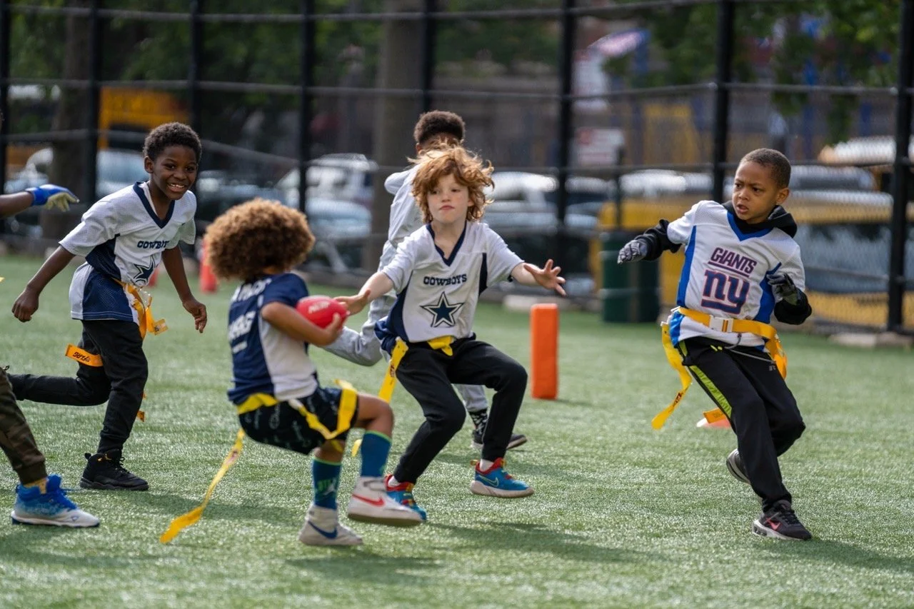 Children playing flag football on a sports field, wearing jerseys with the Dallas Cowboys and New York Giants logos, with a chain-linked fence and parked cars in the background.