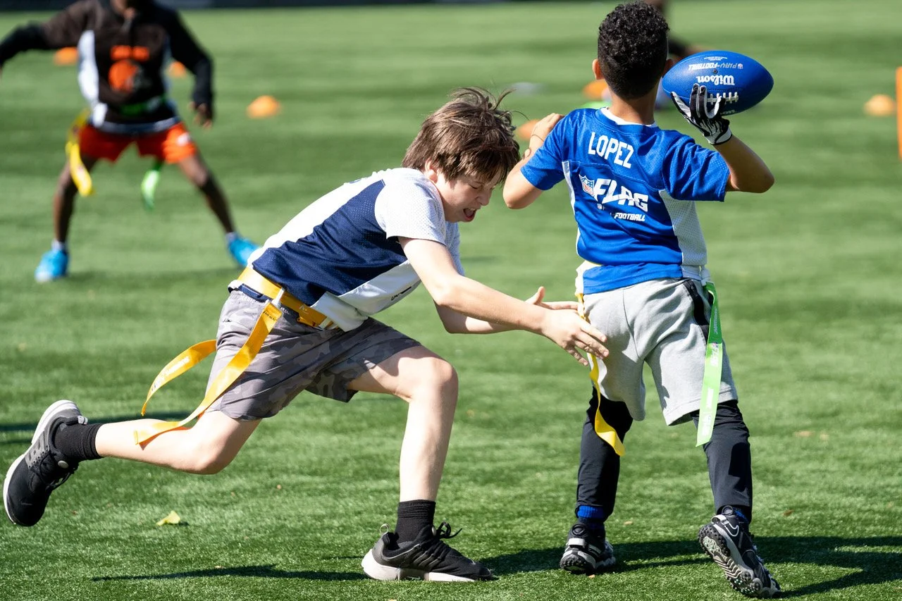 Children playing flag football on a grassy field, one kid in a blue jersey labeled 'Lopez' holding a football, another child attempting to pull the flag from Lopez's waist.