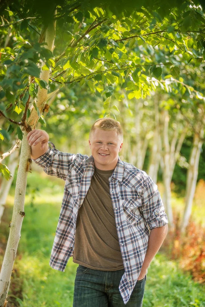 A young man with short blond hair smiling and standing outdoors in a garden or park, leaning against a tree with green leaves.
