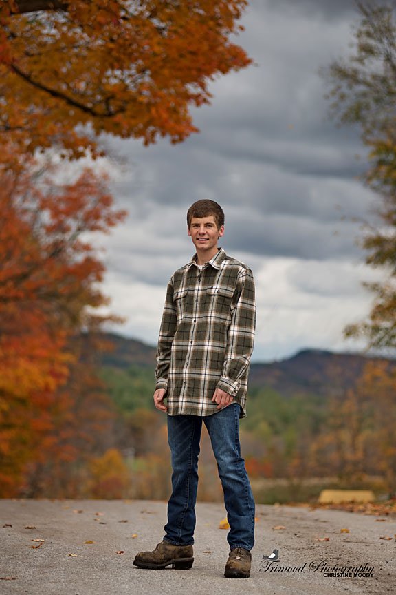 A young man standing outdoors on a paved path surrounded by autumn-colored trees, with mountains in the background.