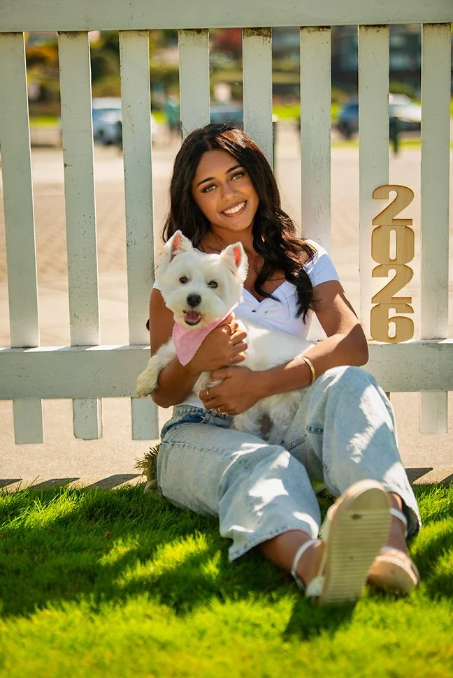 A young woman sitting on the grass, smiling and holding a small white dog with a pink bandana, in front of a white fence with the number 2026. There are cars and trees in the background on a sunny day.