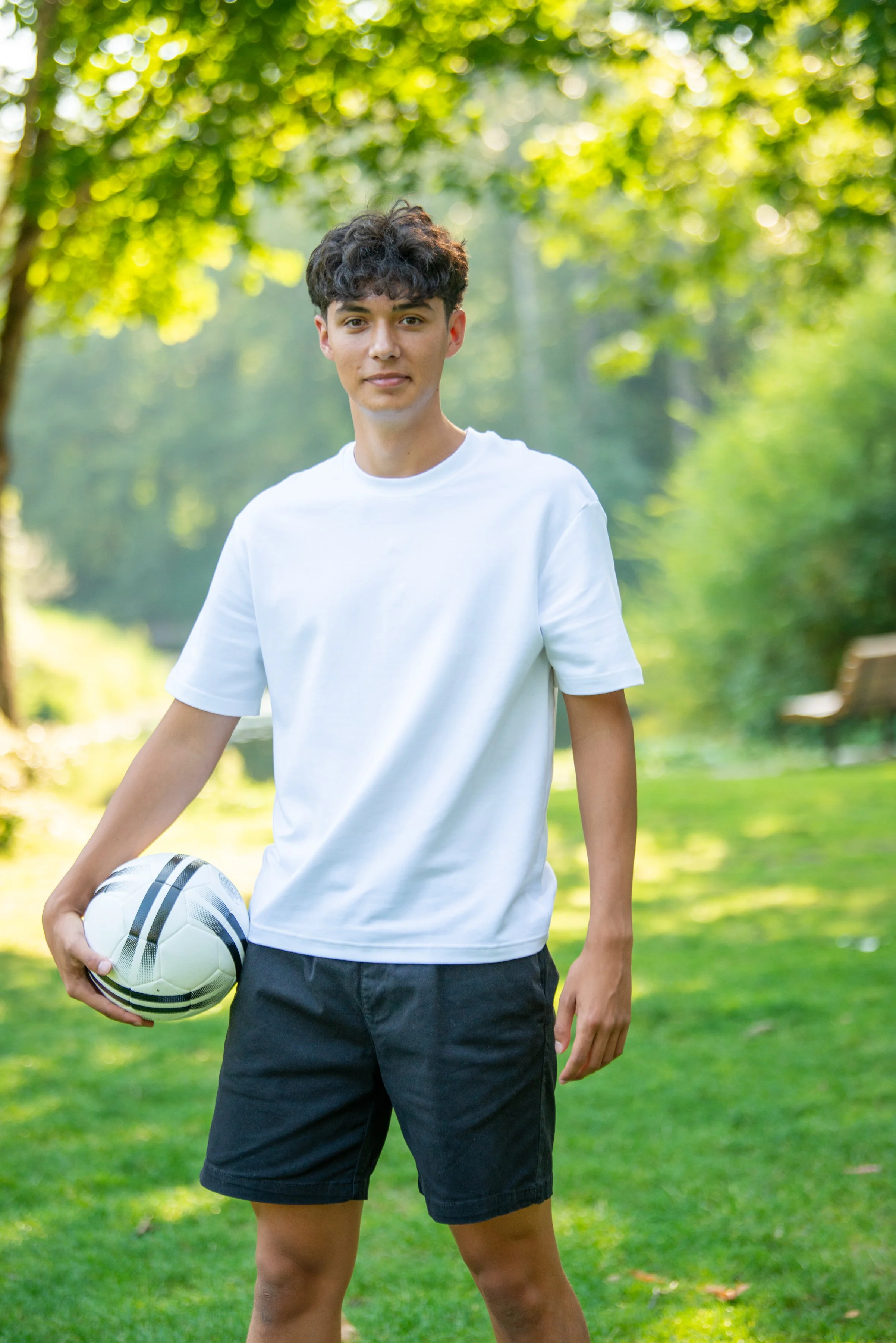 A young man with dark, curly hair is standing outdoors on grass in a park, holding a soccer ball in his left hand. He is wearing a white T-shirt and black shorts, with trees and sunlight in the background.