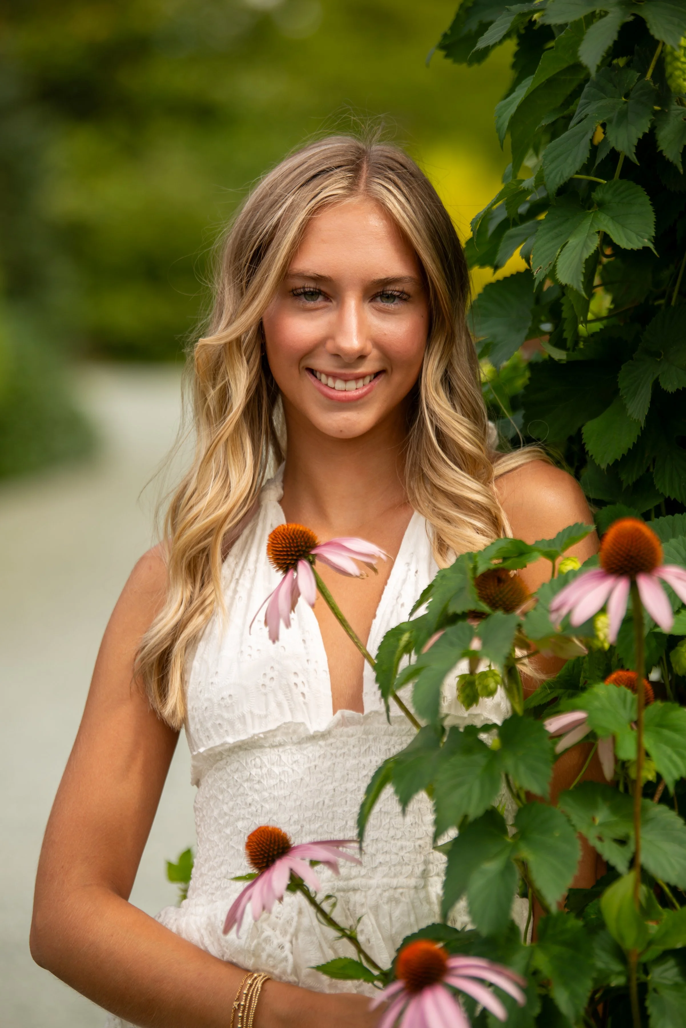 A young woman with long blonde hair, smiling, standing outdoors among green leaves and pink flowers.