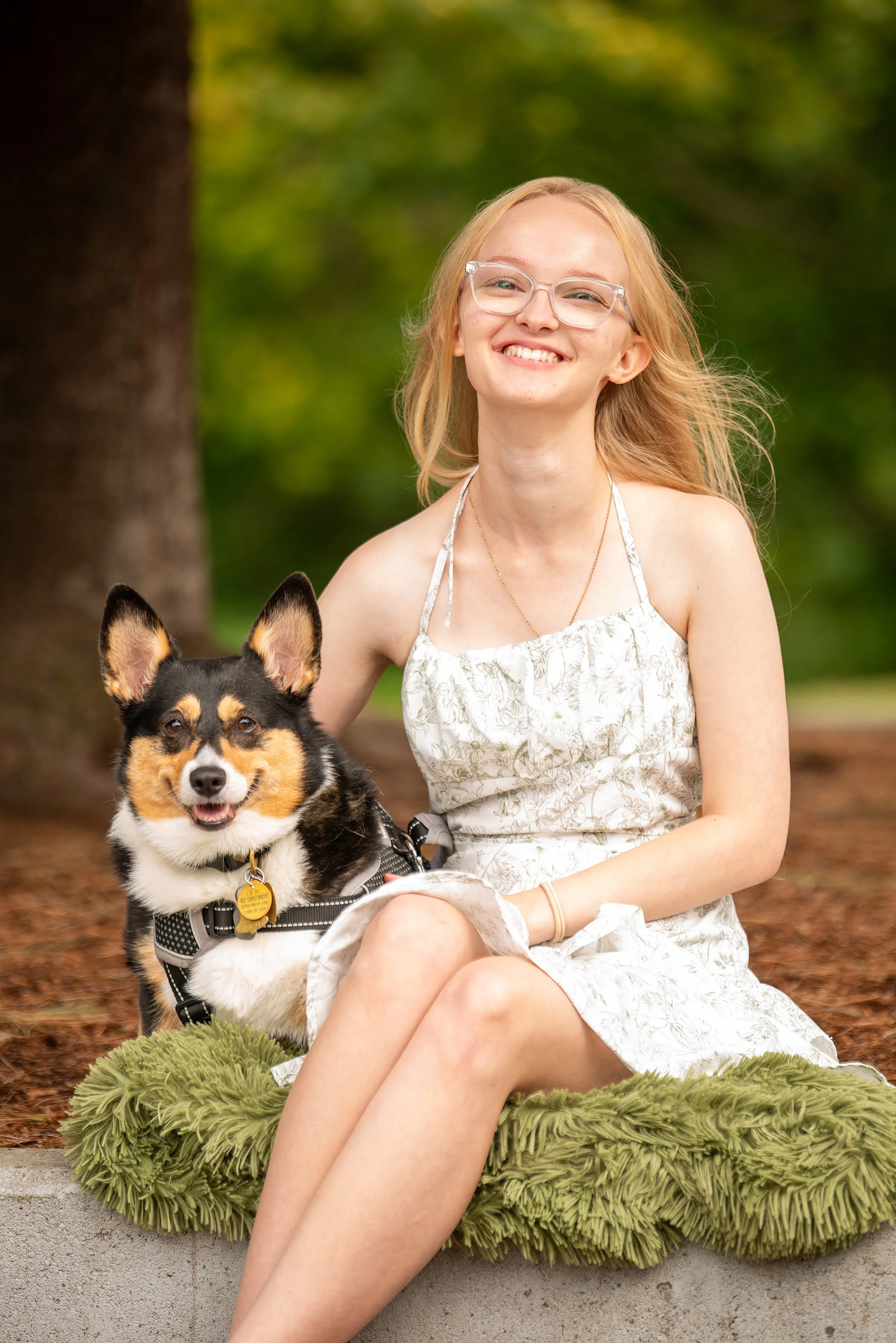 A young woman with blonde hair, glasses, and a white floral dress sitting on a green fuzzy cushion in an outdoor park, smiling, with a black and tan dog sitting beside her.