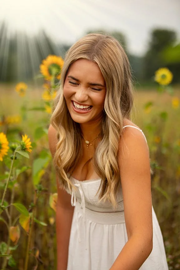 A young woman with long blonde hair wearing a white dress, smiling and laughing in a sunflower field during daytime.