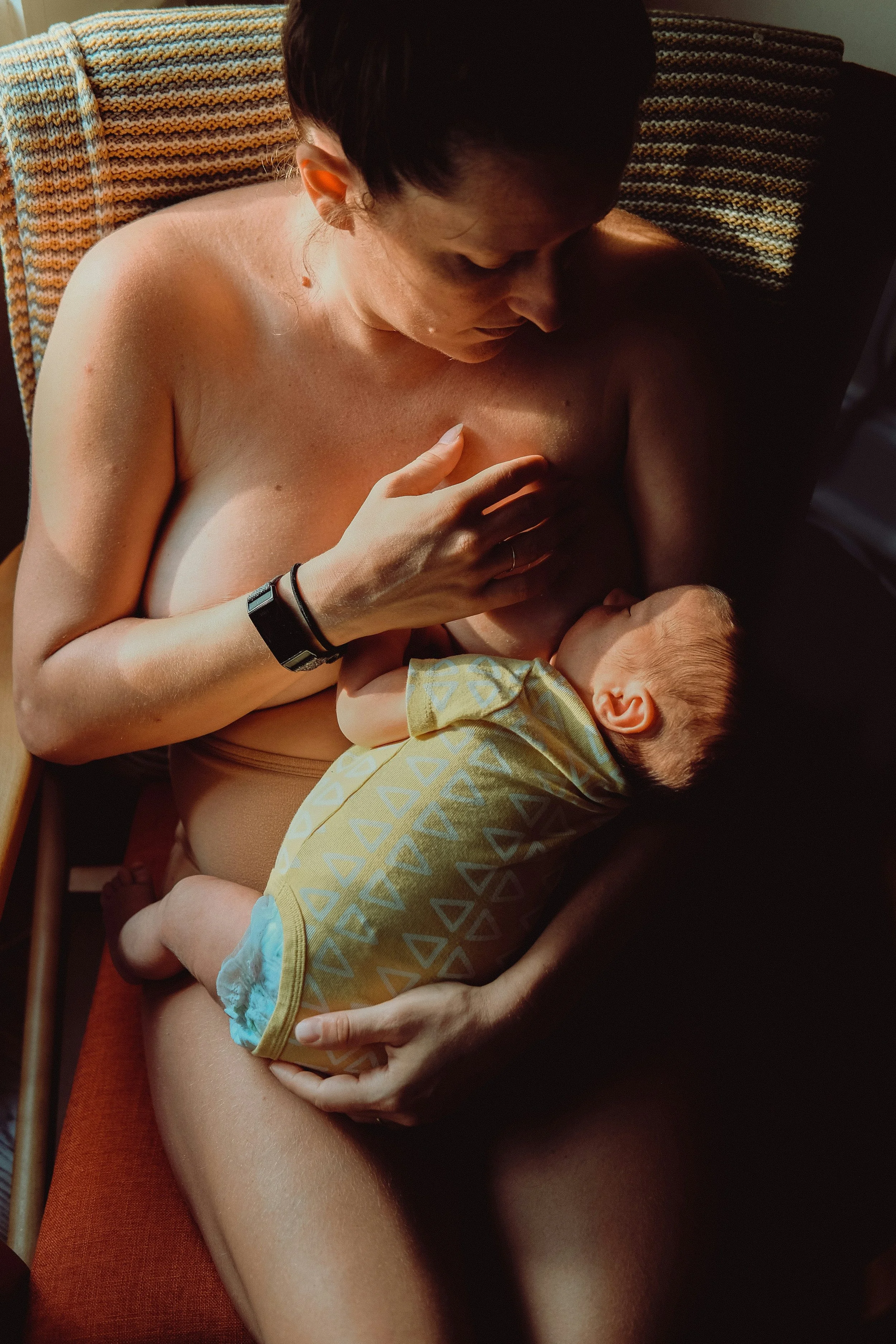 A woman breastfeeding a baby on a reddish-brown chair with a knitted colorful blanket behind her.