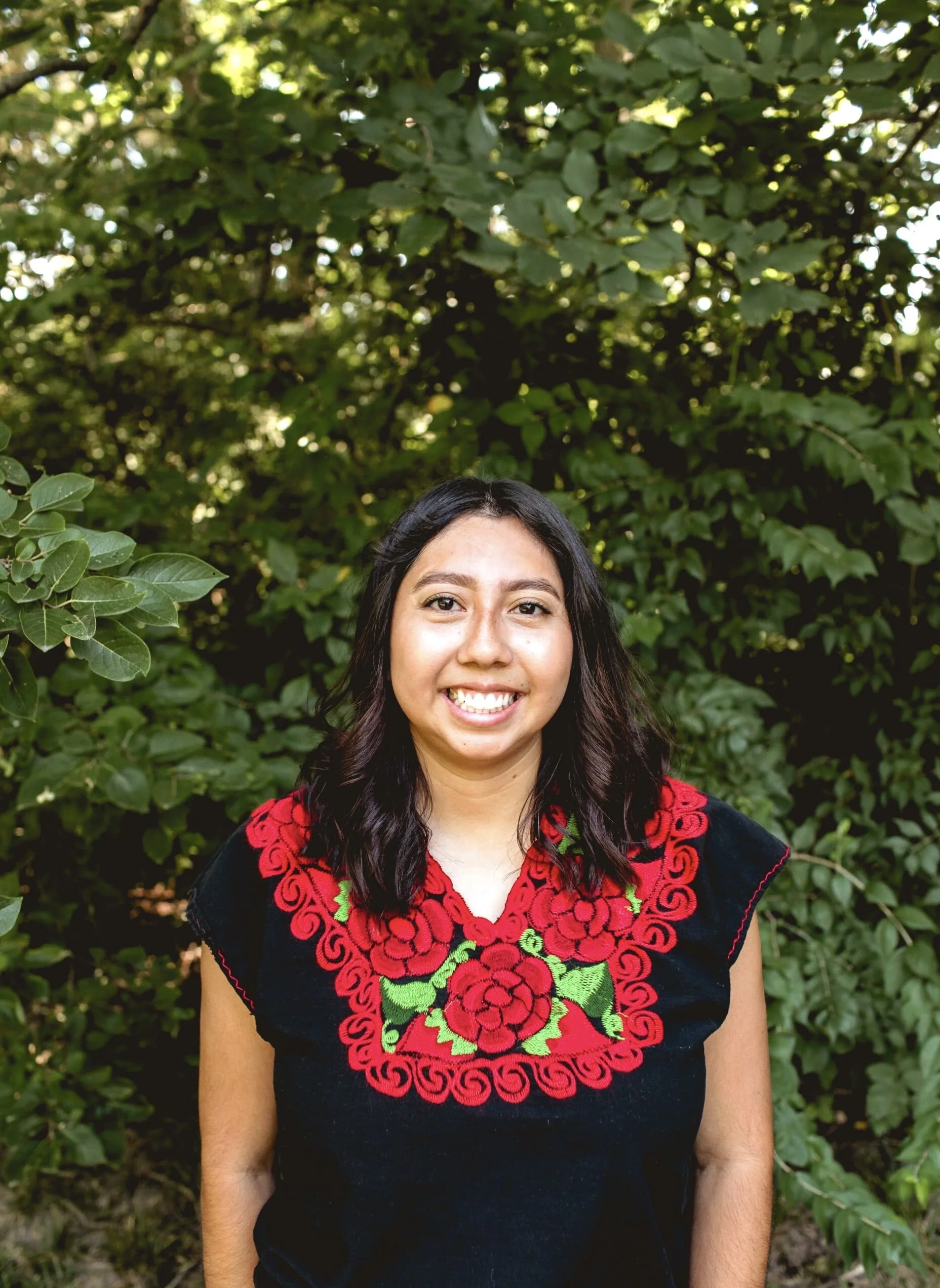 A woman with long black hair and a bright smile, wearing a traditional black garment with red and green embroidery, standing outdoors in front of lush green foliage.