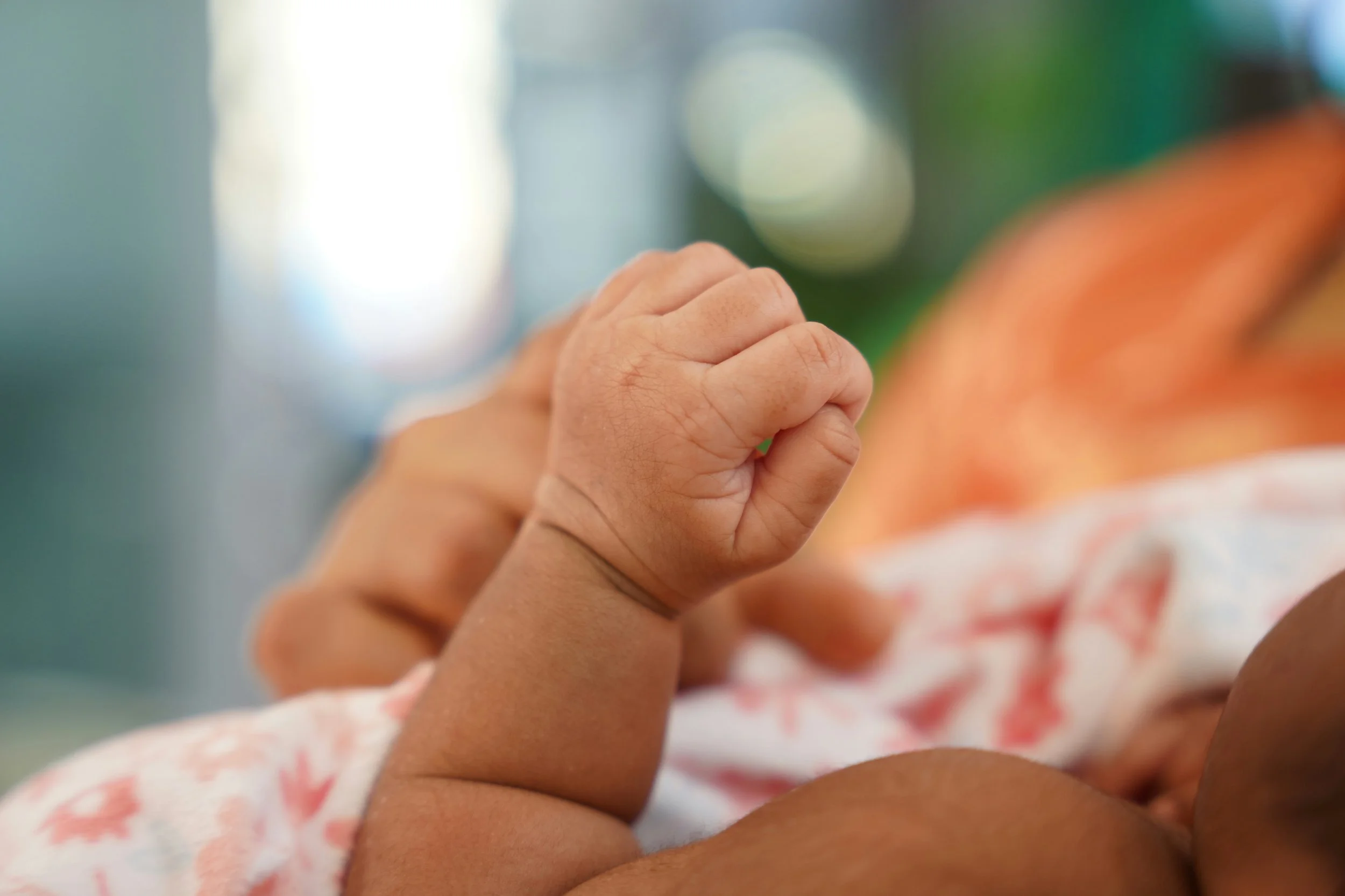 Close-up of a baby's hand clasped around an adult's finger, with a blurred background of colorful patterns.