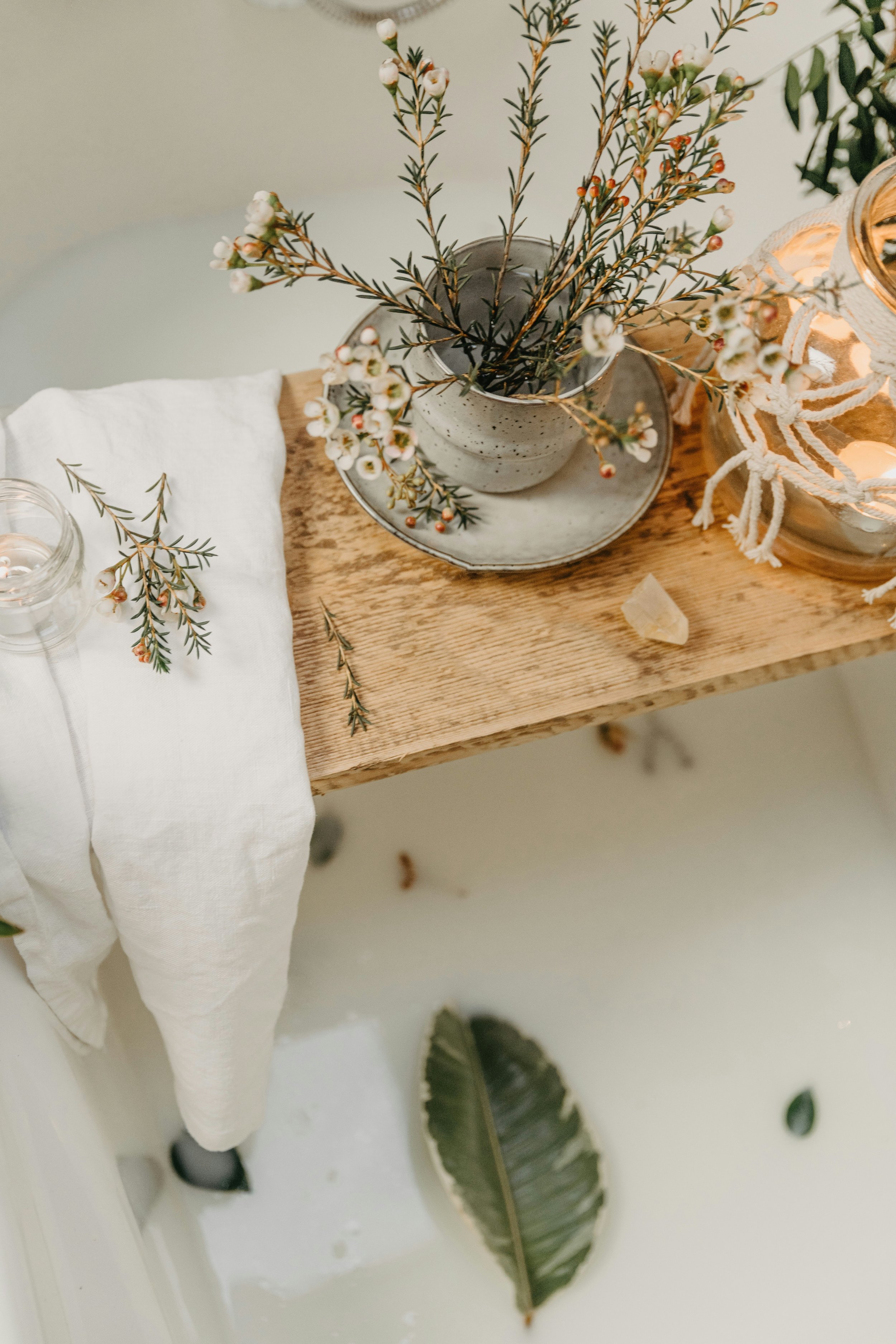 Top-down view of a wooden table with a potted plant, a candle in a jar, a white cloth, and a small piece of rock. There are scattered sprigs of green leaves and a large leaf on the floor.