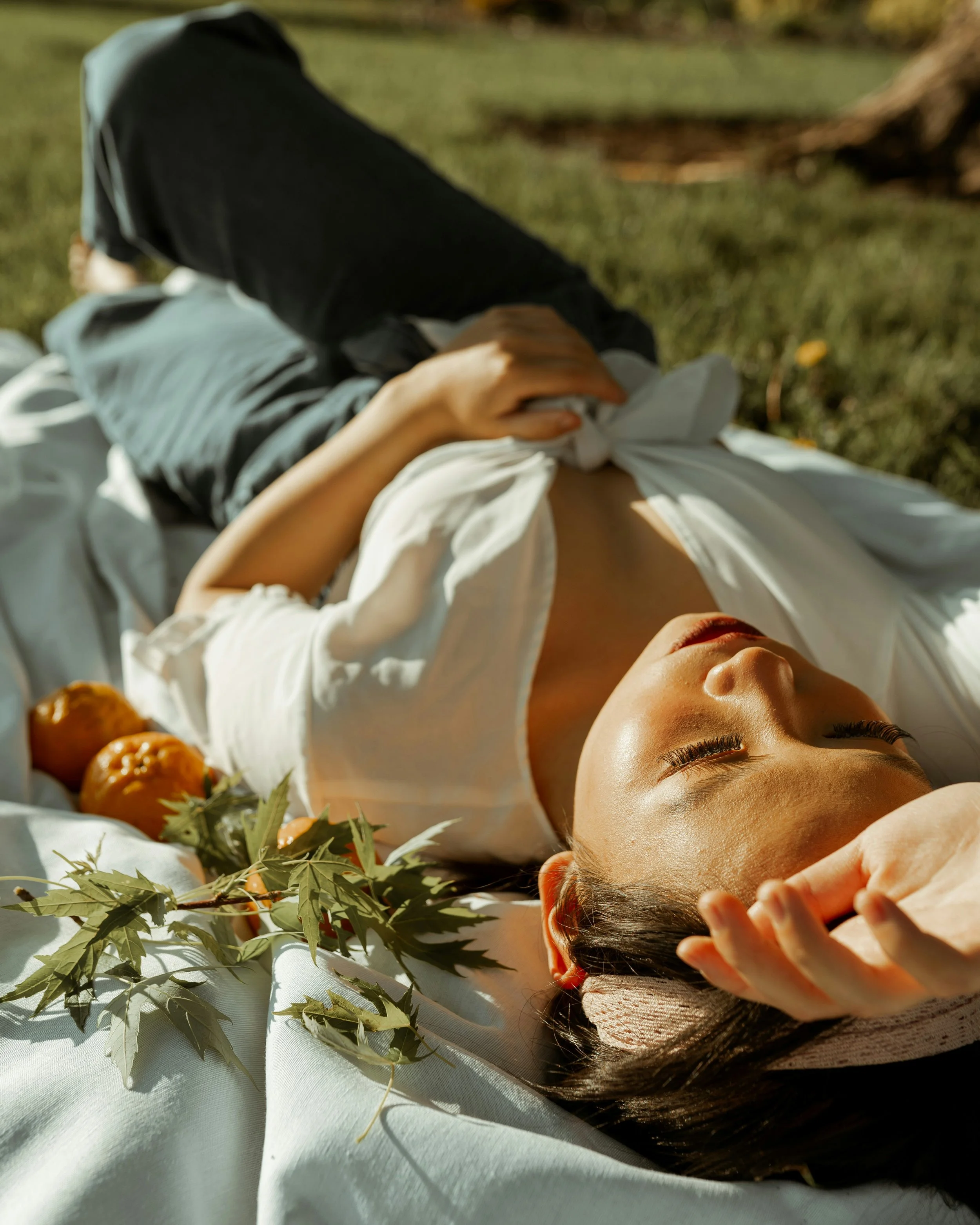 A woman lying down on a white surface outdoors, with green leaves and orange fruit nearby, in a peaceful scene.