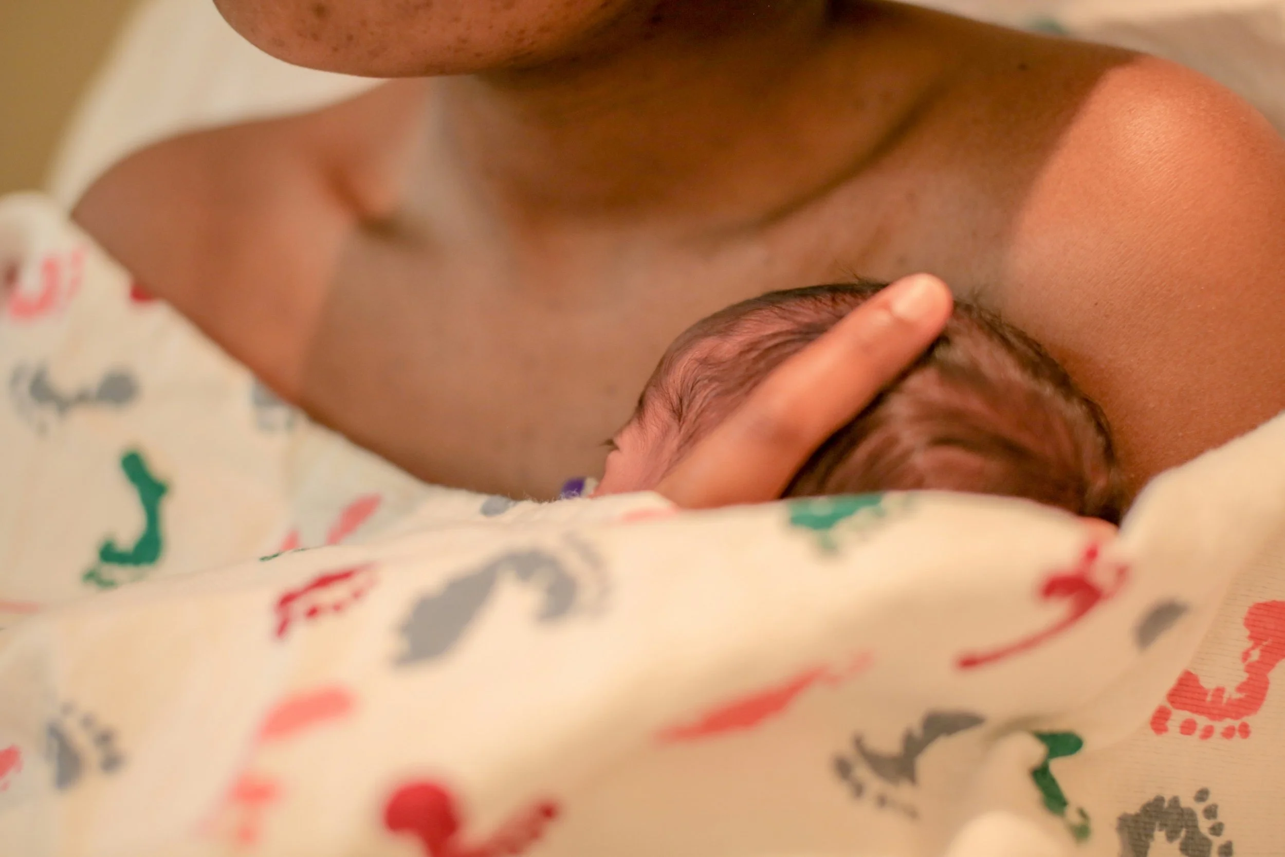Close-up of a newborn baby lying on a blanket with colorful dinosaur patterns, gently holding an adult's finger.