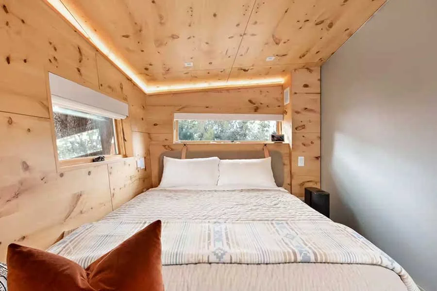 A cozy bedroom with wooden walls and ceiling, a bed with white pillows and a beige patterned blanket, small windows on the side and headboard, and a black speaker next to the bed.
